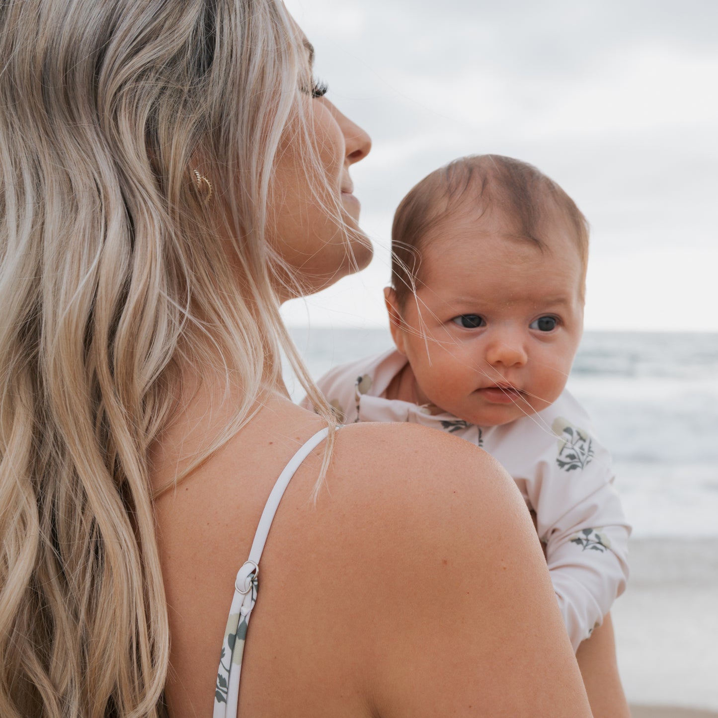 A woman with long blonde hair stands on a beach, holding a baby in her arms. The baby wears the forever french baby Girl's Long Sleeve Swimsuit in Marigold and gazes calmly ahead as the ocean extends behind them.