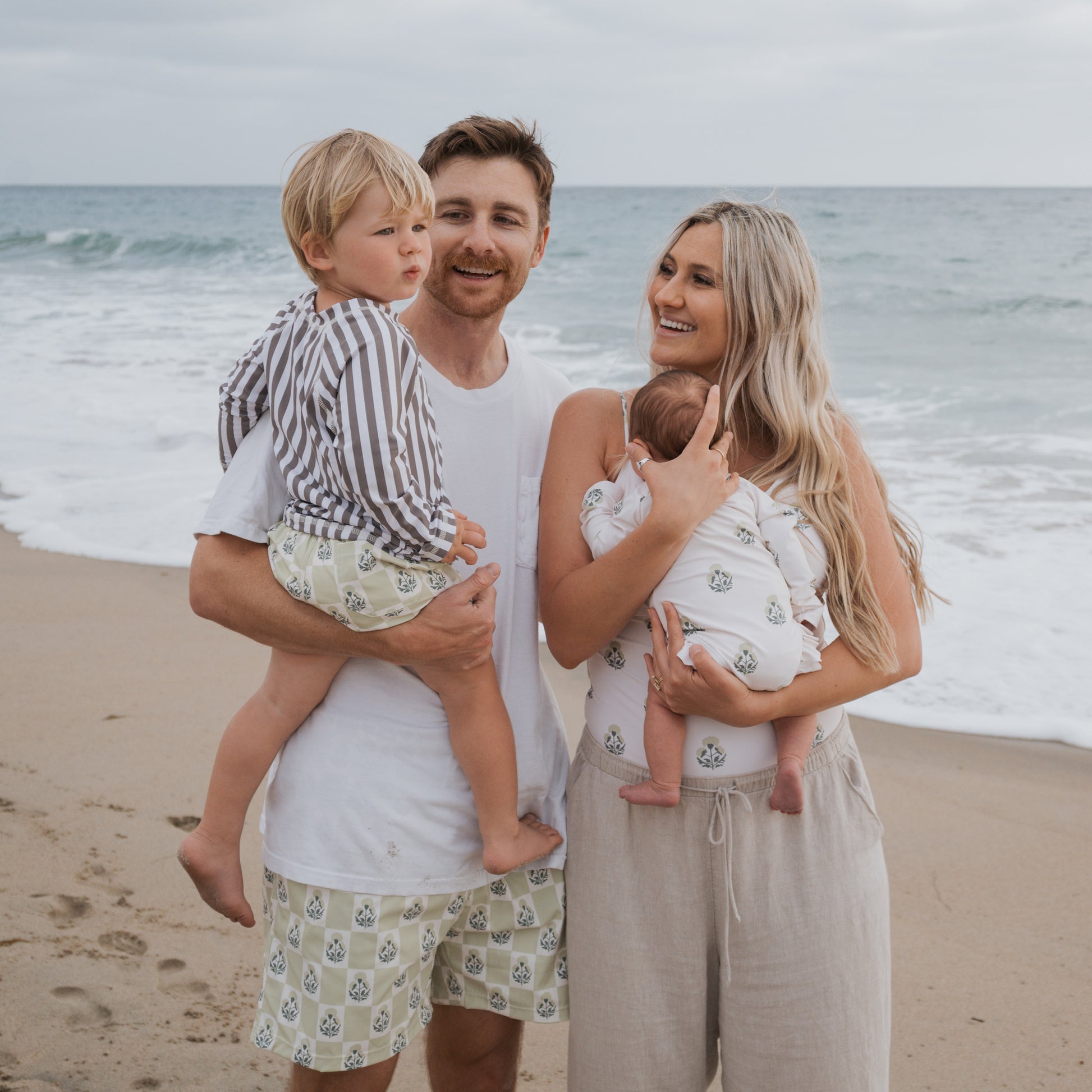 A smiling family of four stands on the beach near the ocean. The young boy wears the forever french baby Child Board Short in Marigold Check, while everyone else is dressed in light, casual summer clothes under an overcast sky.