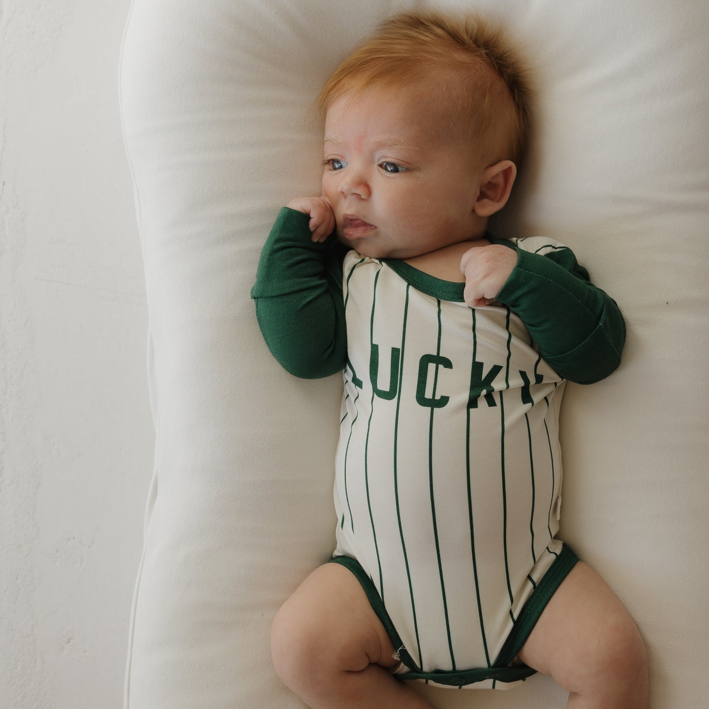 A light-skinned baby with reddish hair lies on a white cushion, wearing the forever french baby Long Sleeve Bamboo Snapsuit | Lucky, featuring cream and green stripes with "LUCKY" on the front. Their hands are near their mouth as they look to the side.