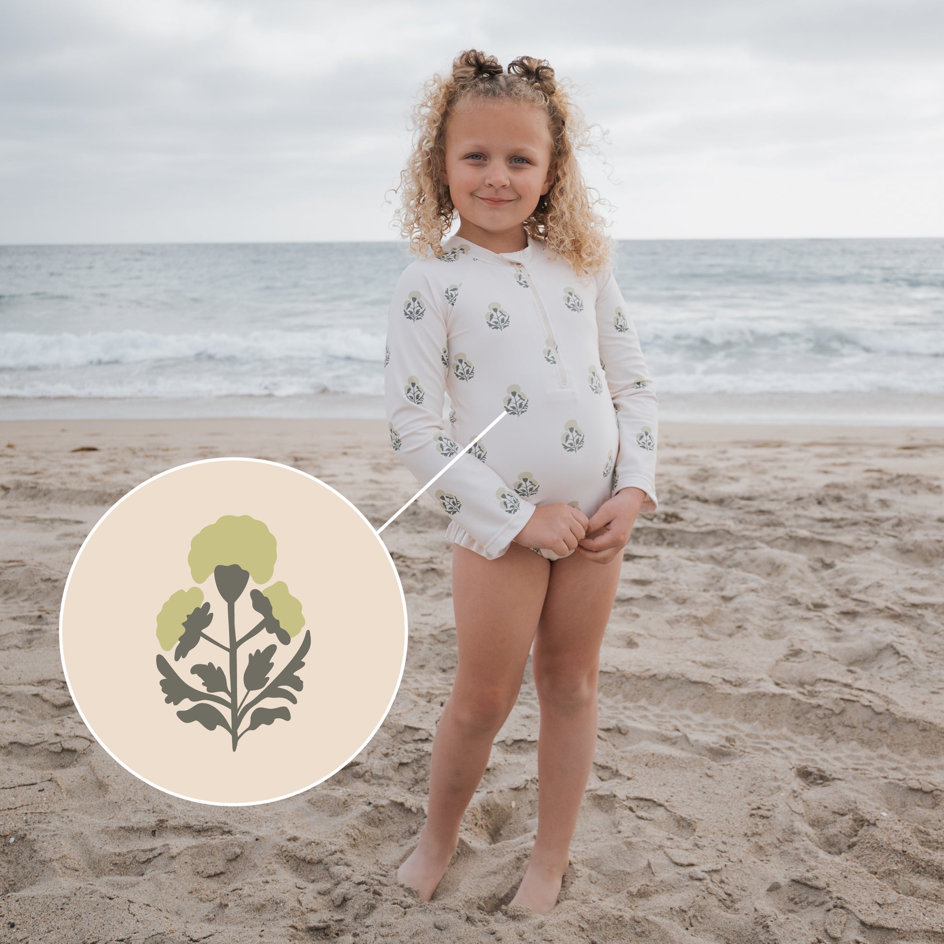 A young child with curly hair stands on a sandy beach near the ocean, wearing the forever french baby Girl's Long Sleeve Swimsuit | Marigold, featuring a green floral pattern with ruffle detail. A close-up highlights the green and gray flower print.