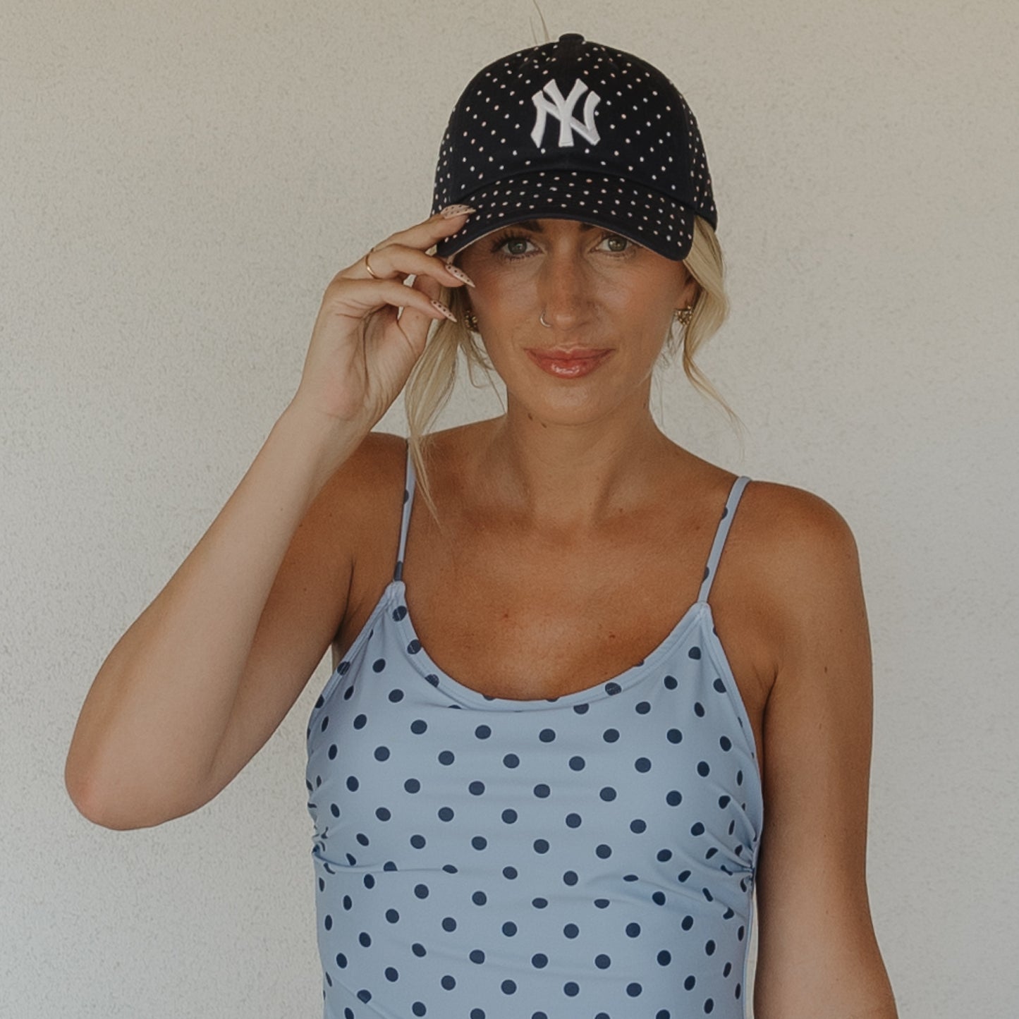 A woman in the Forever French Women's One Piece Swimsuit | Sully and a black polka dot Yankees cap stands against a light background, holding her cap and looking at the camera—chic summer beachwear inspiration.