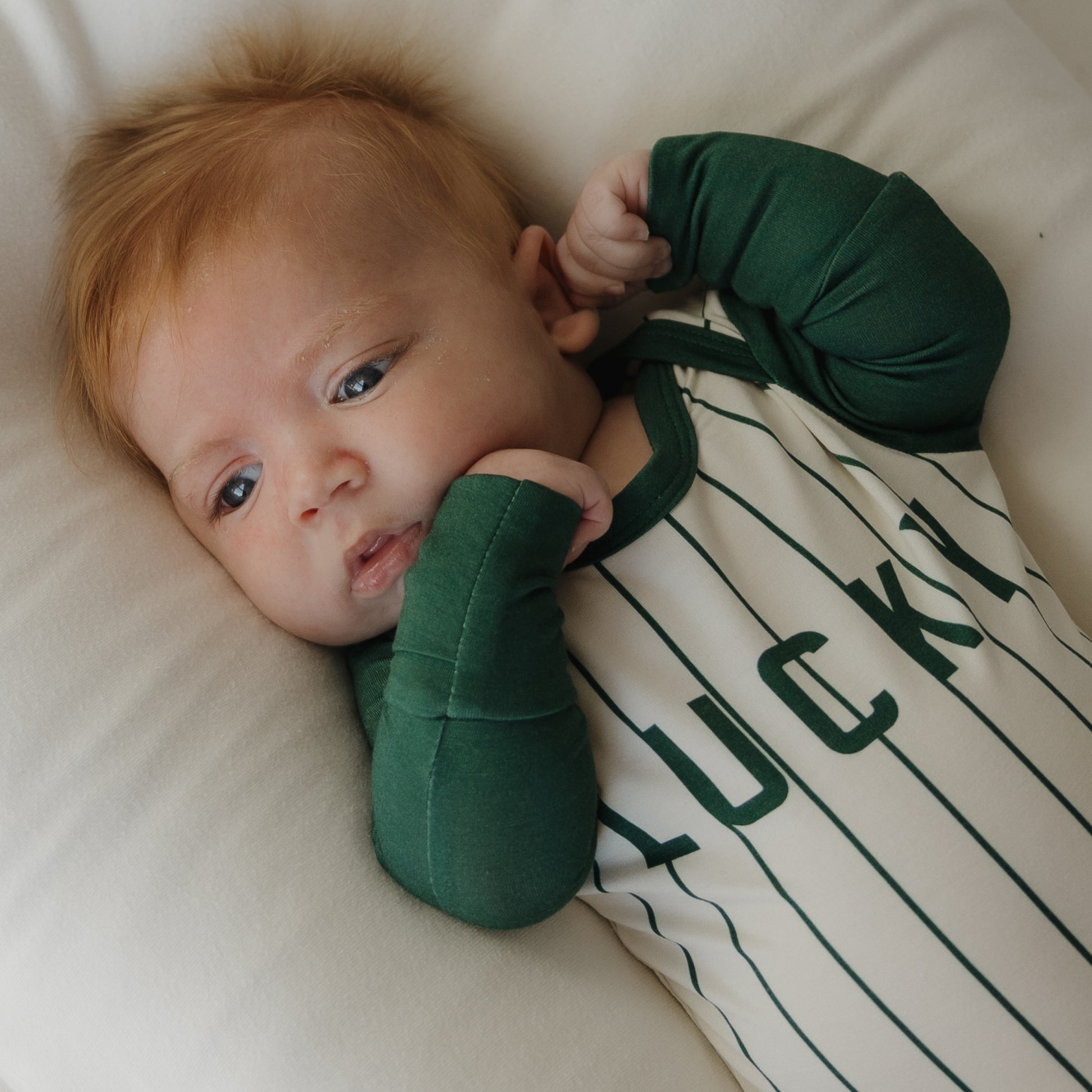 A red-haired baby lies on a white pillow wearing the "Long Sleeve Bamboo Snapsuit | Lucky" by forever french baby, featuring green sleeves, striped design, and "LUCKY" on the front for stylish comfort in gentle bamboo fabric.