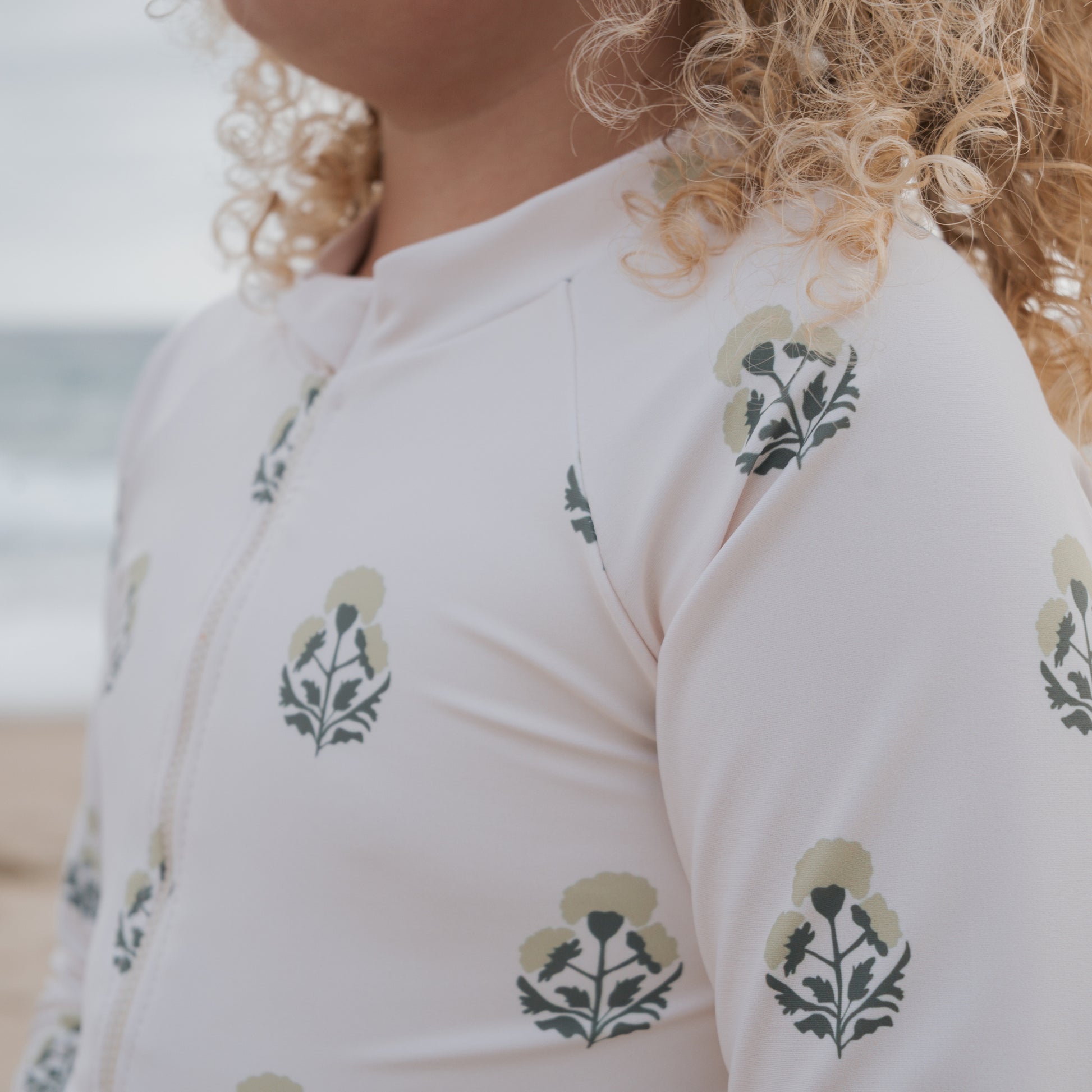 A blonde child stands on the beach before the ocean, wearing the forever french baby Girl's Long Sleeve Swimsuit in Marigold, a light-colored one-piece with a floral print.