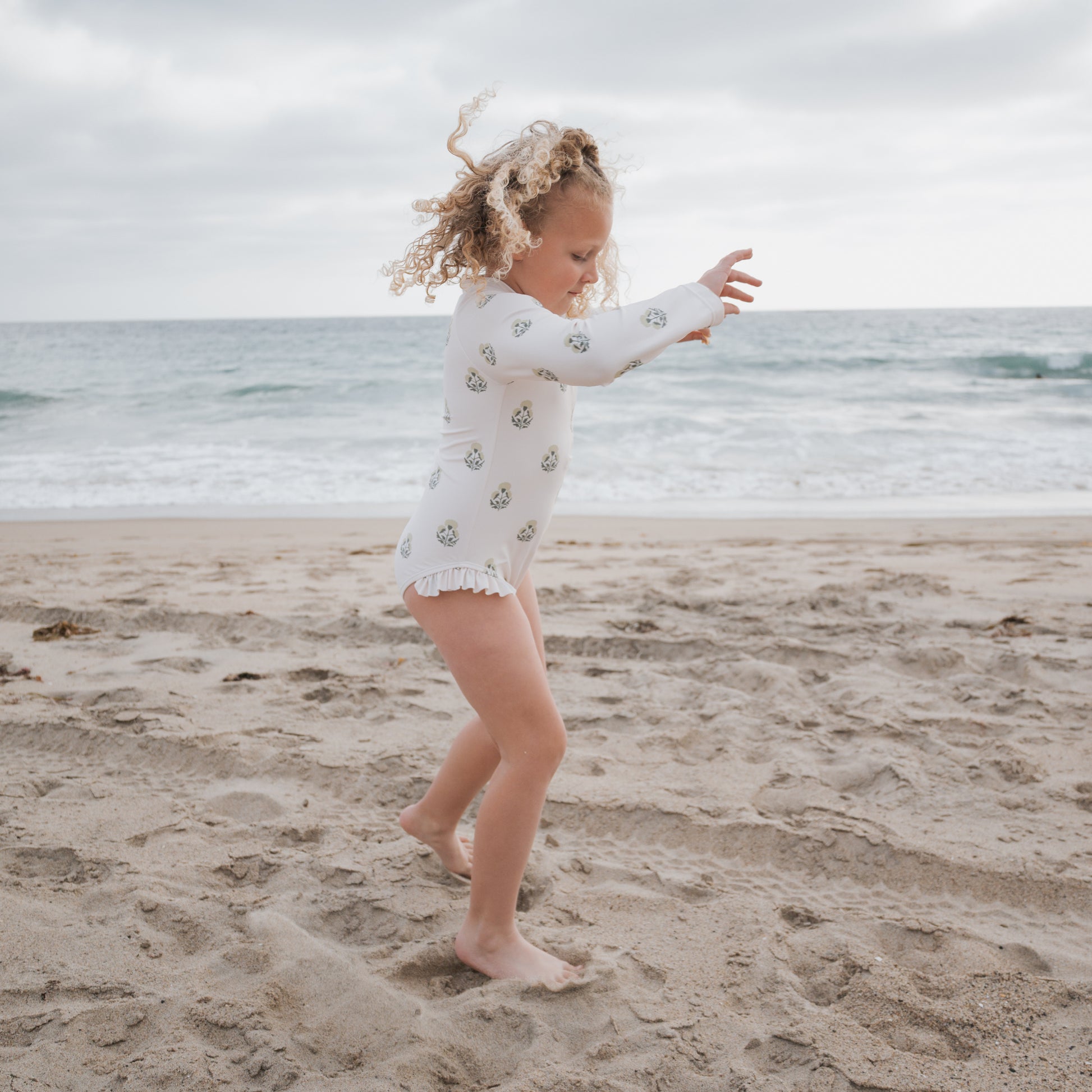 A young girl in the forever french baby Girl's Long Sleeve Swimsuit | Marigold walks barefoot on a sandy beach, playing as waves roll in beneath a cloudy sky.