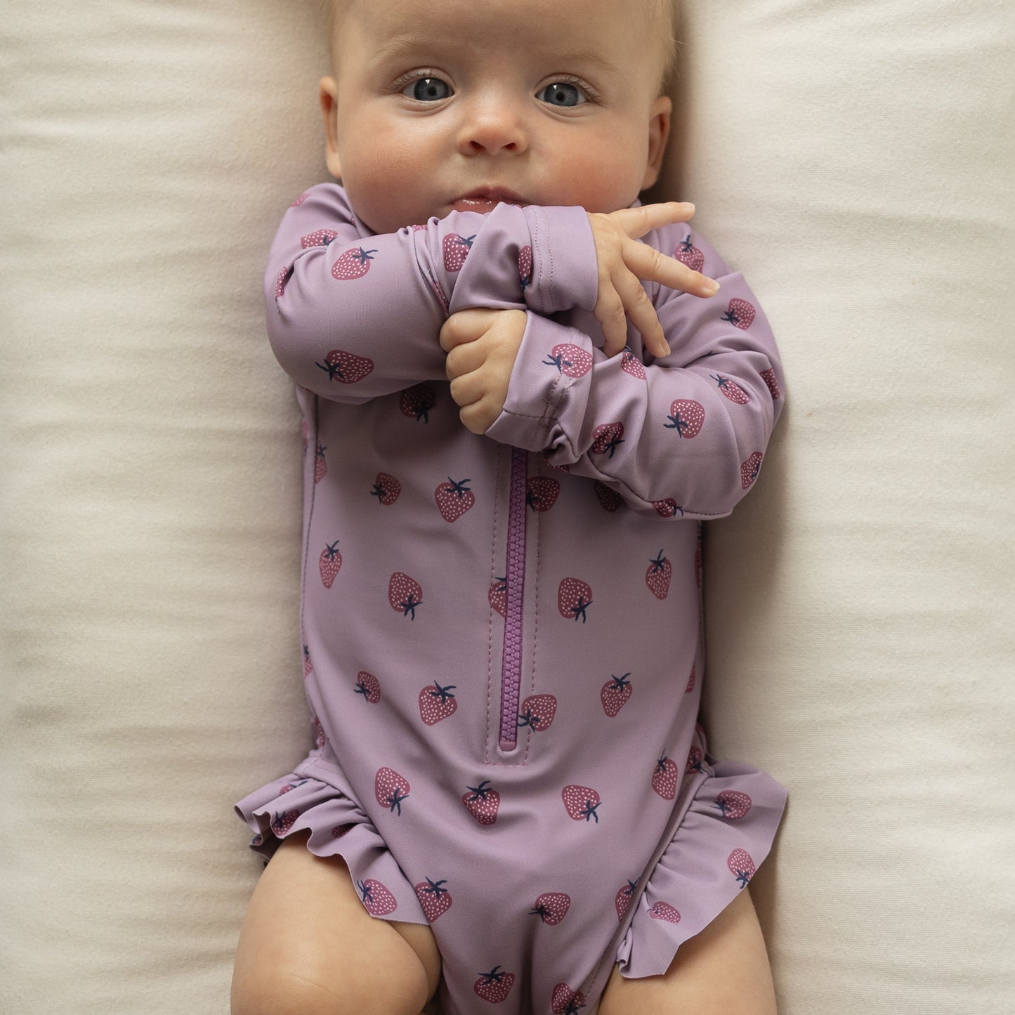 A baby wearing the forever french baby Girl's Long Sleeve Swimsuit | Matilda lies on a cream-colored blanket, looking up with arms crossed near their face.