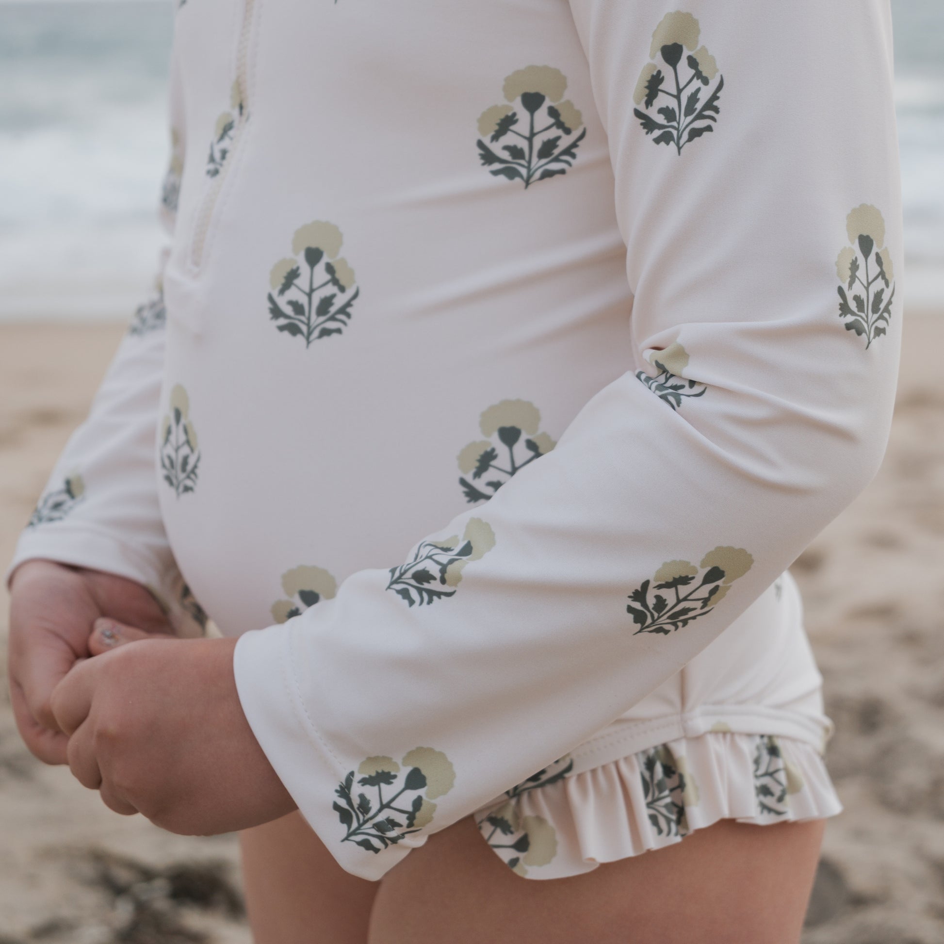 A child in the forever french baby Girl's Long Sleeve Swimsuit | Marigold, featuring a dandelion print and ruffle details, stands on a sandy beach near the water with hands clasped at their waist.
