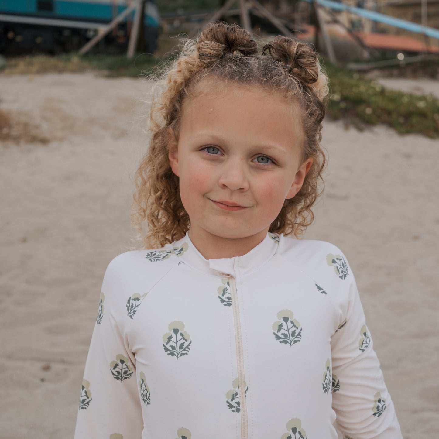 A young girl with curly blonde hair styled in two buns stands on a sandy beach, wearing the forever french baby Girl's Long Sleeve Swimsuit in Marigold.