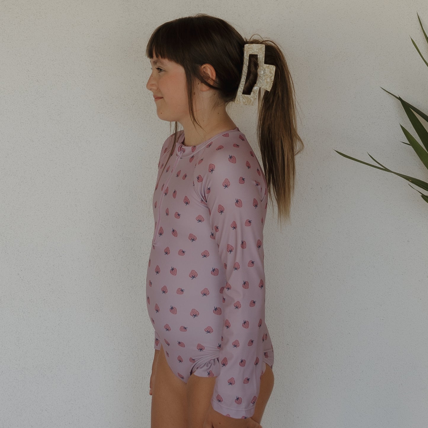 A young girl with long brown hair in a ponytail, secured by a large clip, stands in profile wearing the forever french baby Girl's Long Sleeve Swimsuit | Matilda in light purple with pink shells, against a white wall with a plant on the right.