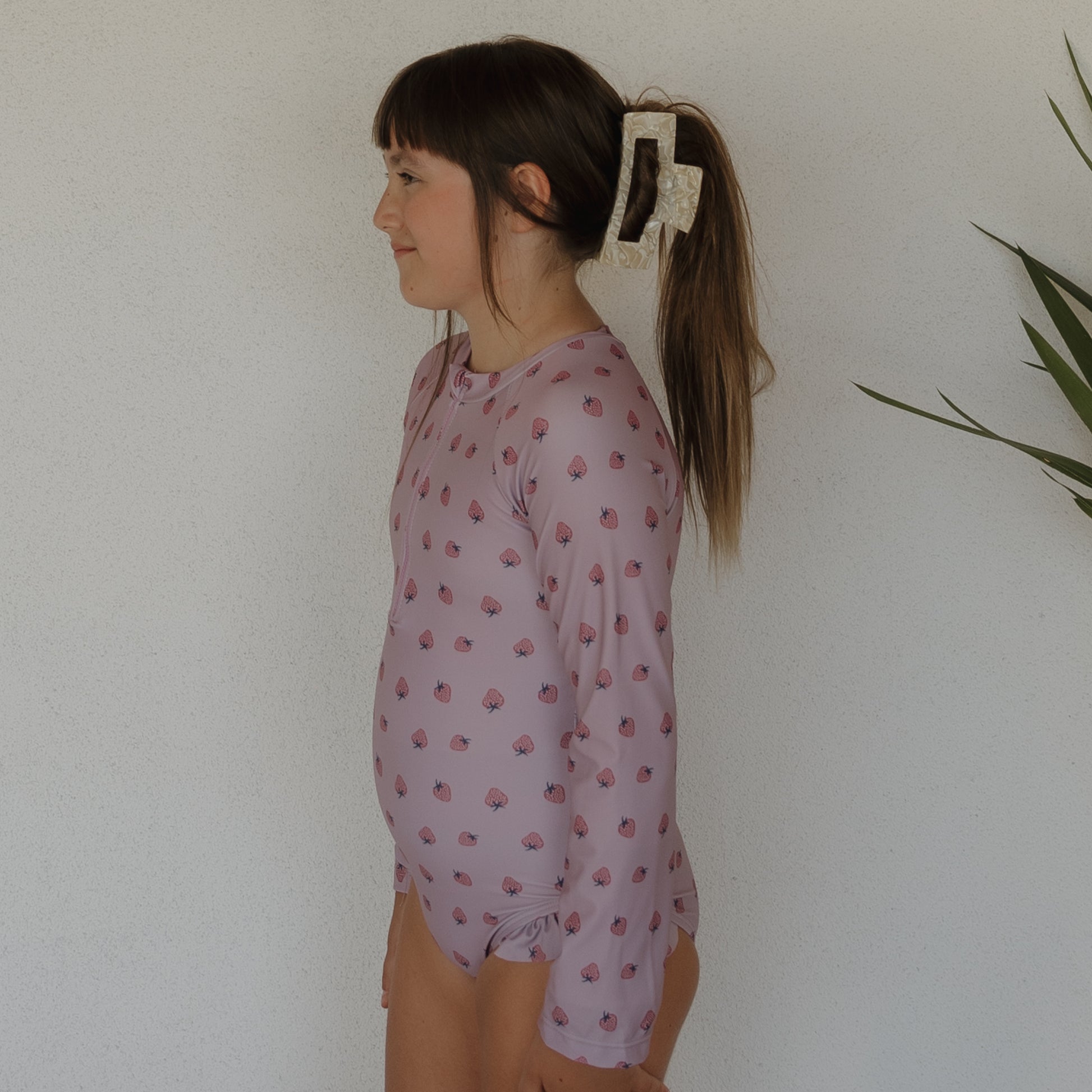 A young girl with long brown hair in a ponytail, secured by a large clip, stands in profile wearing the forever french baby Girl's Long Sleeve Swimsuit | Matilda in light purple with pink shells, against a white wall with a plant on the right.