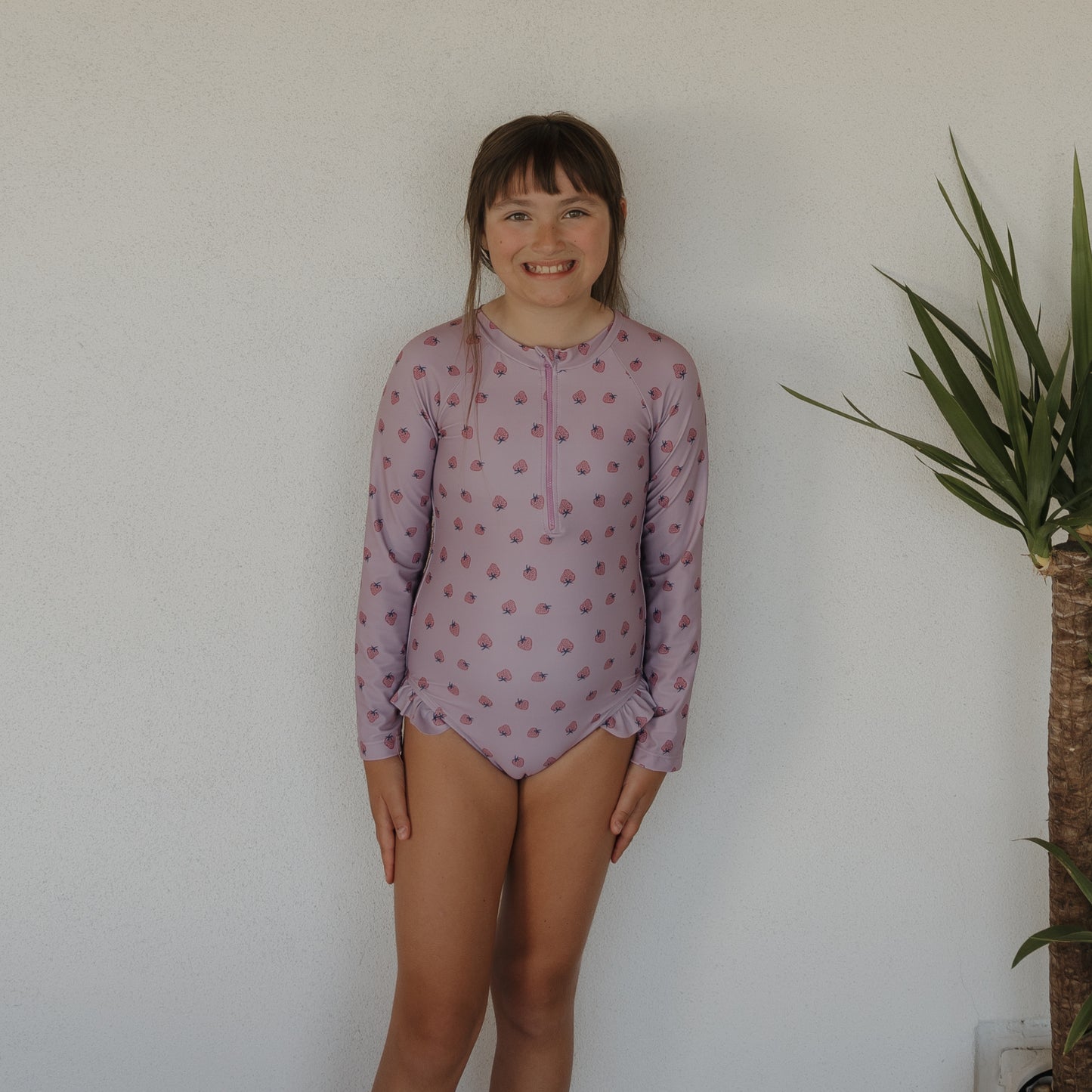 A young girl smiles against a white wall, wearing the forever french baby Girl's Long Sleeve Swimsuit | Matilda in lavender with a small print pattern. A green potted plant is visible to her right.