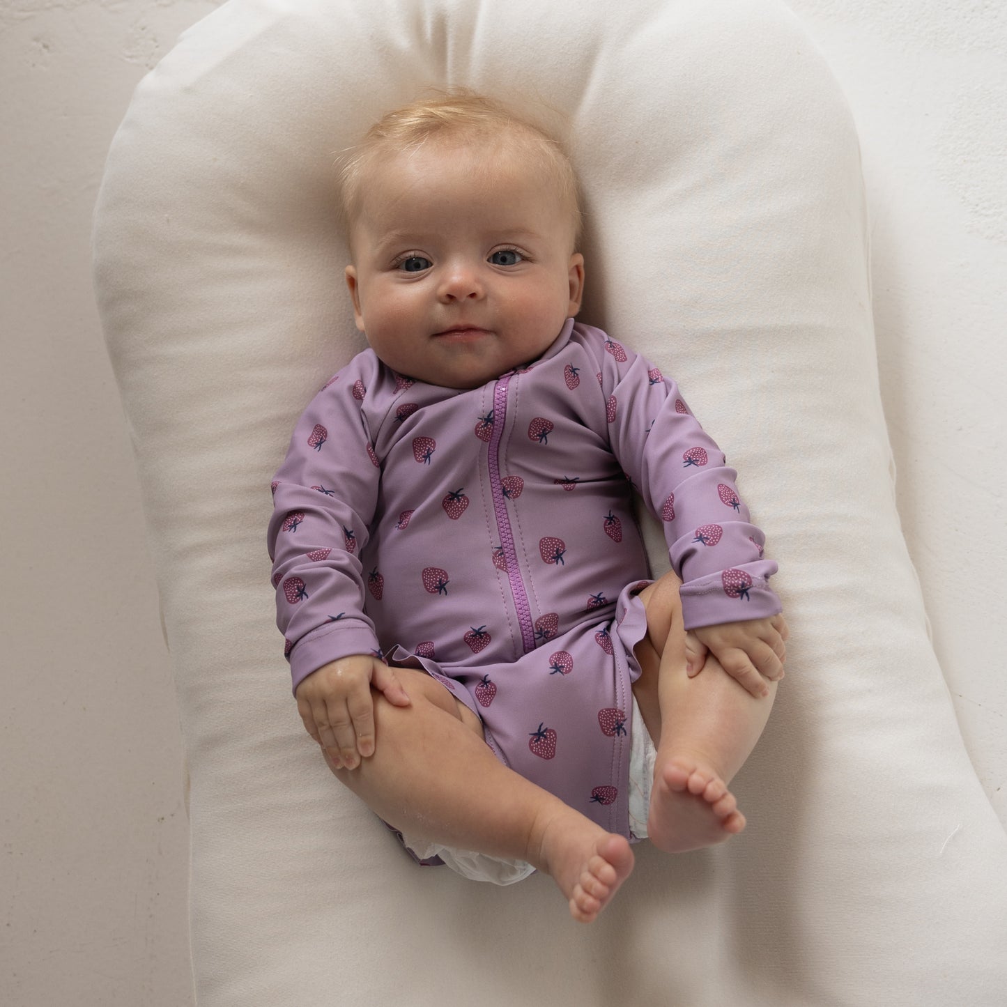A light-haired baby wears the forever french baby Girl's Long Sleeve Swimsuit | Matilda, featuring a purple and pink pattern, while lying on a soft white cushion and looking gently at the camera.