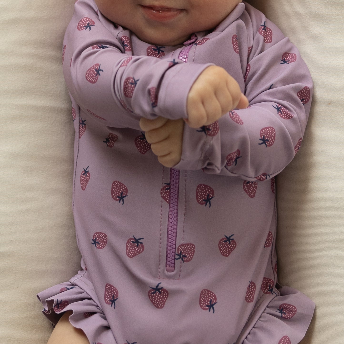 A baby smiles while lying on a light surface, wearing the forever french baby Girl's Long Sleeve Swimsuit | Matilda featuring a purple color, strawberry pattern, and ruffles, with hands playfully crossed in front of their chest.