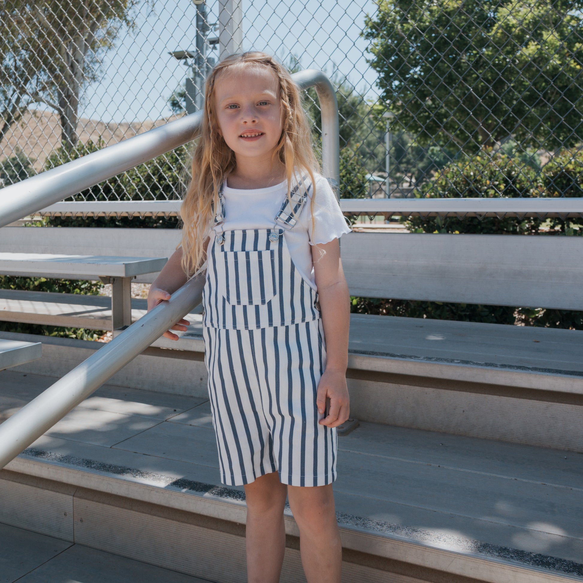 A young girl with long blonde hair stands on bleachers, holding a metal railing. She wears a white shirt and forever french baby Overalls | Denim Stripe. Trees and a wire fence are seen in the sunny background.