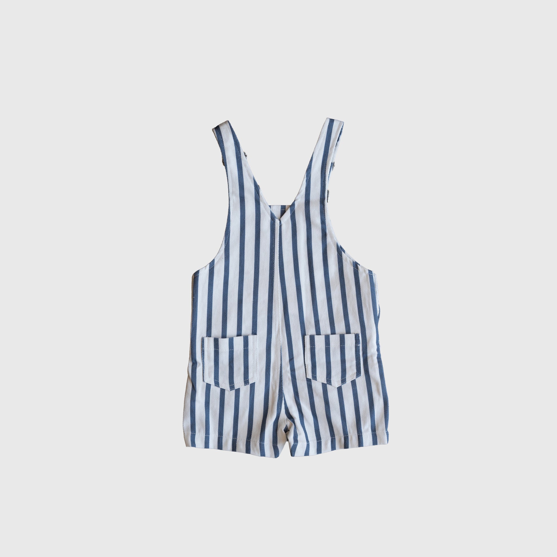 The Overalls | Denim Stripe by forever french baby is a blue and white vertically striped sleeveless romper with two front pockets, shown laid flat on a light gray background.