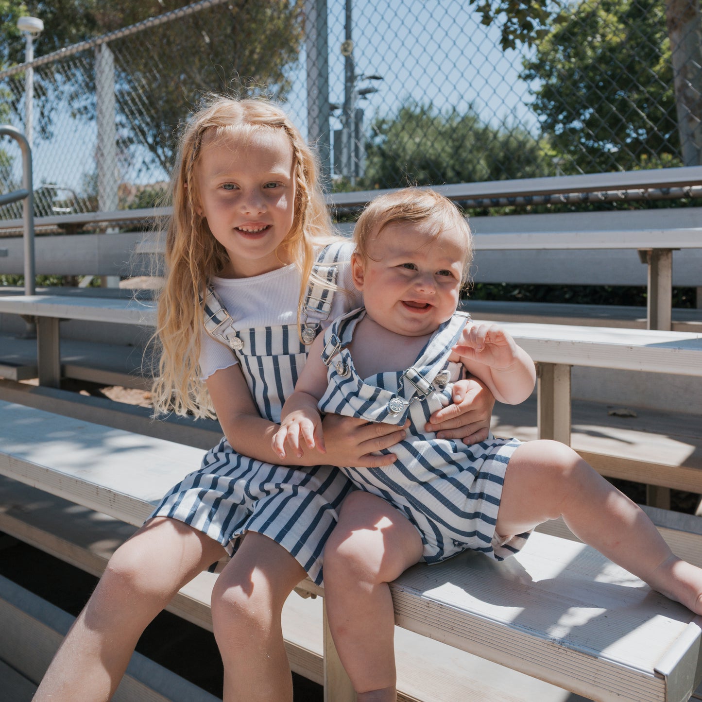 Two young children sit outdoors on bleachers. The older child smiles while holding a baby; both wear matching "Denim Stripe" overalls from forever french baby. Sunlight gently filters through, with a fence and trees seen in the background.