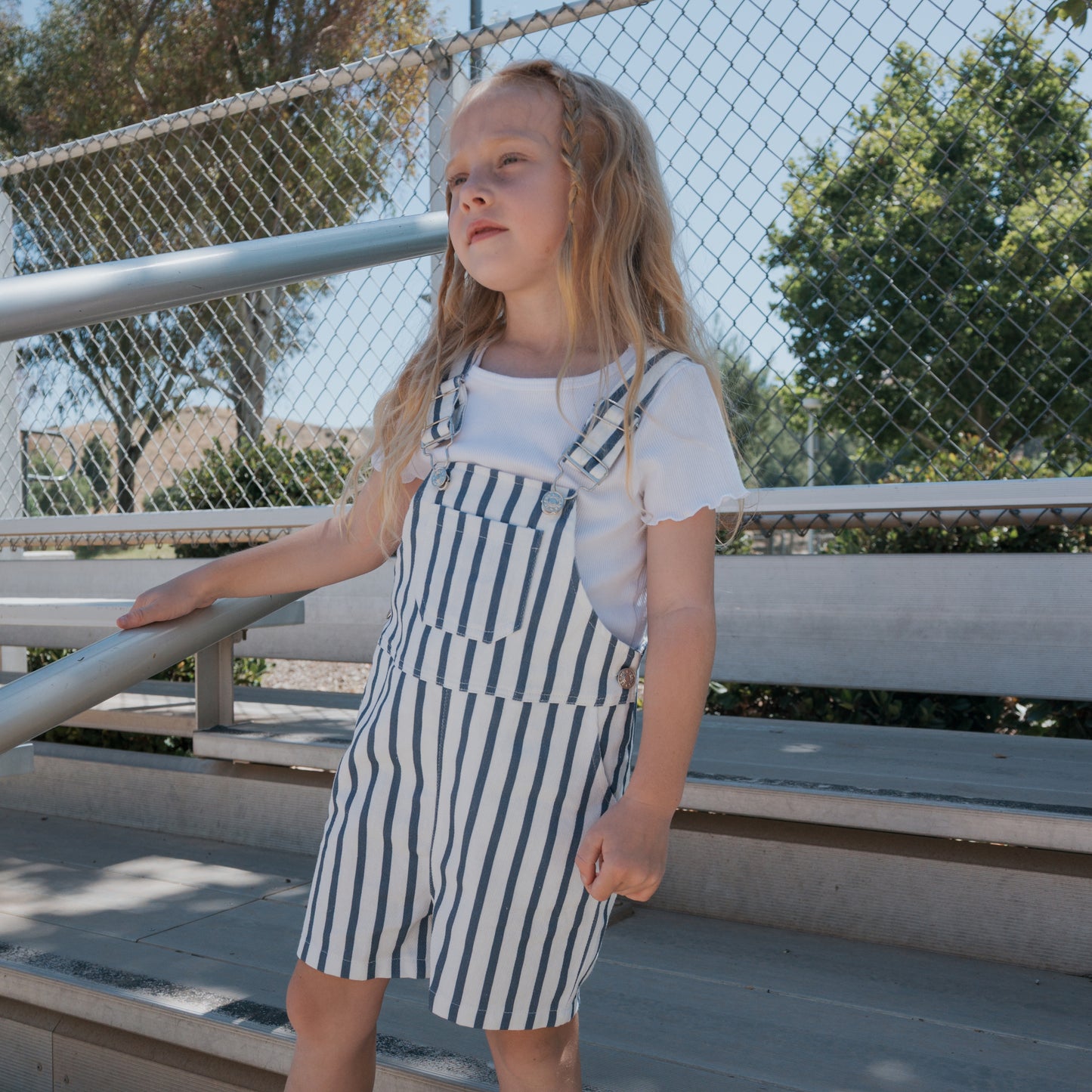 A young girl wearing a white t-shirt and forever french baby Overalls | Denim Stripe stands on metal bleachers near a chain-link fence, with trees and blue sky in the background.