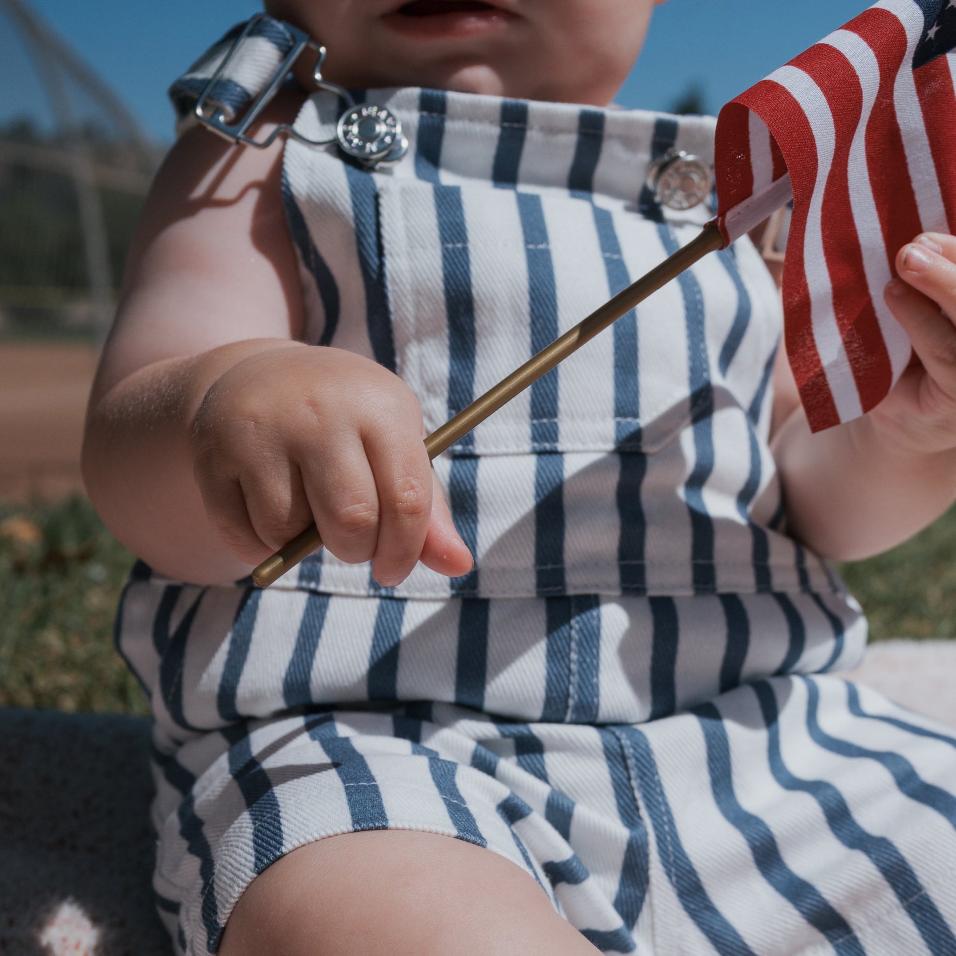 A young child wearing forever french baby Overalls | Denim Stripe sits outdoors in bright sunlight, holding a small American flag. The child's face is partially visible, focusing on their hands and stylish denim outfit.