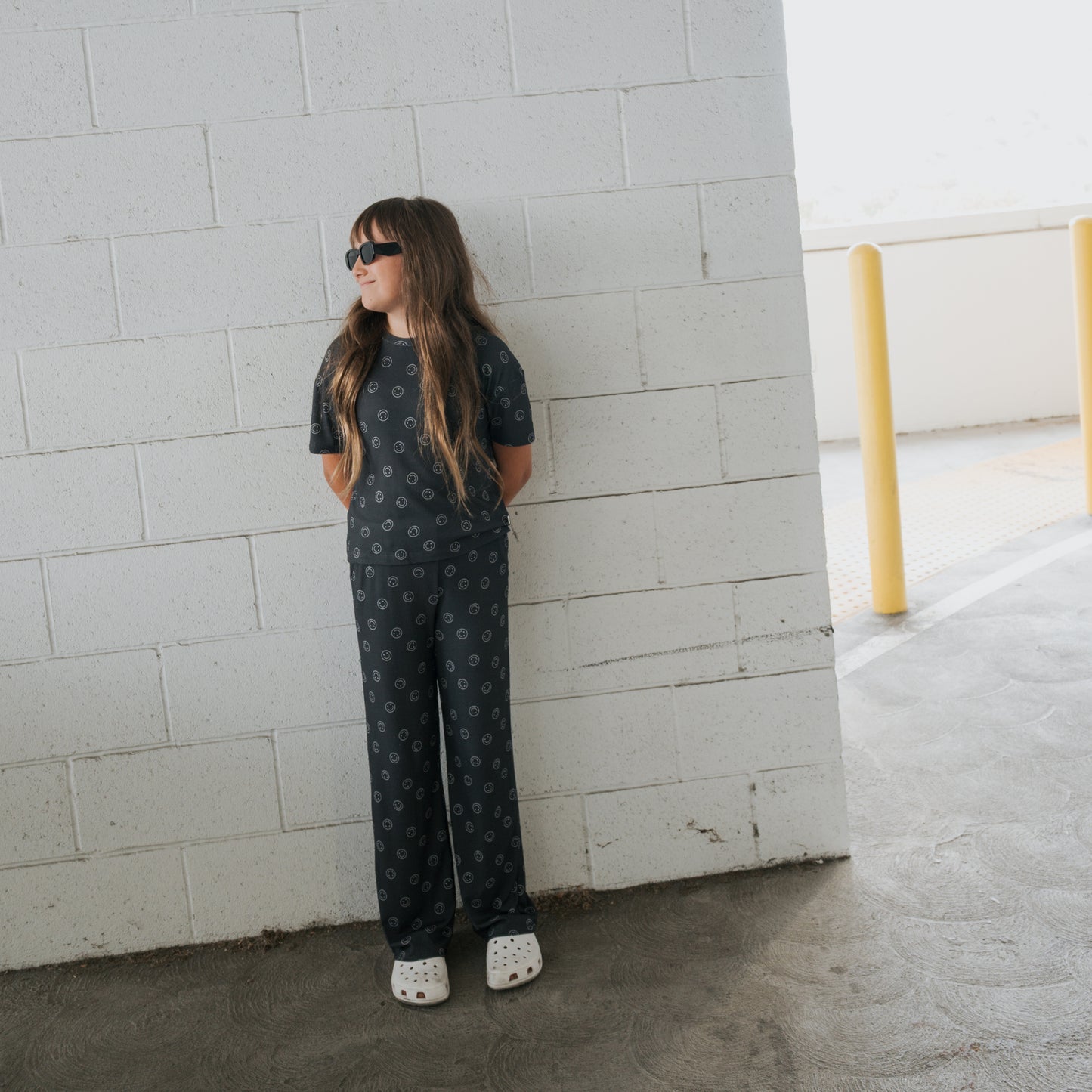 A girl wears the forever french baby Child Boxy Tee Lounge Set in Smiley Ribbed, paired with white Crocs and sunglasses, standing against a white brick wall with her hands behind her back, looking to the side in a parking area.