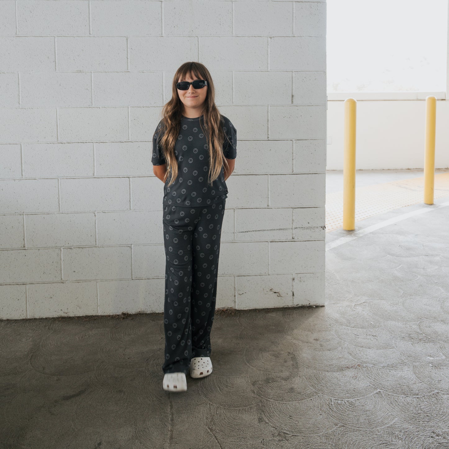 A young person in the forever french baby Child Boxy Tee Lounge Set | Smiley Ribbed stands in front of a white brick wall in a parking garage, sporting long hair, black sunglasses, and white clogs with hands behind their back.