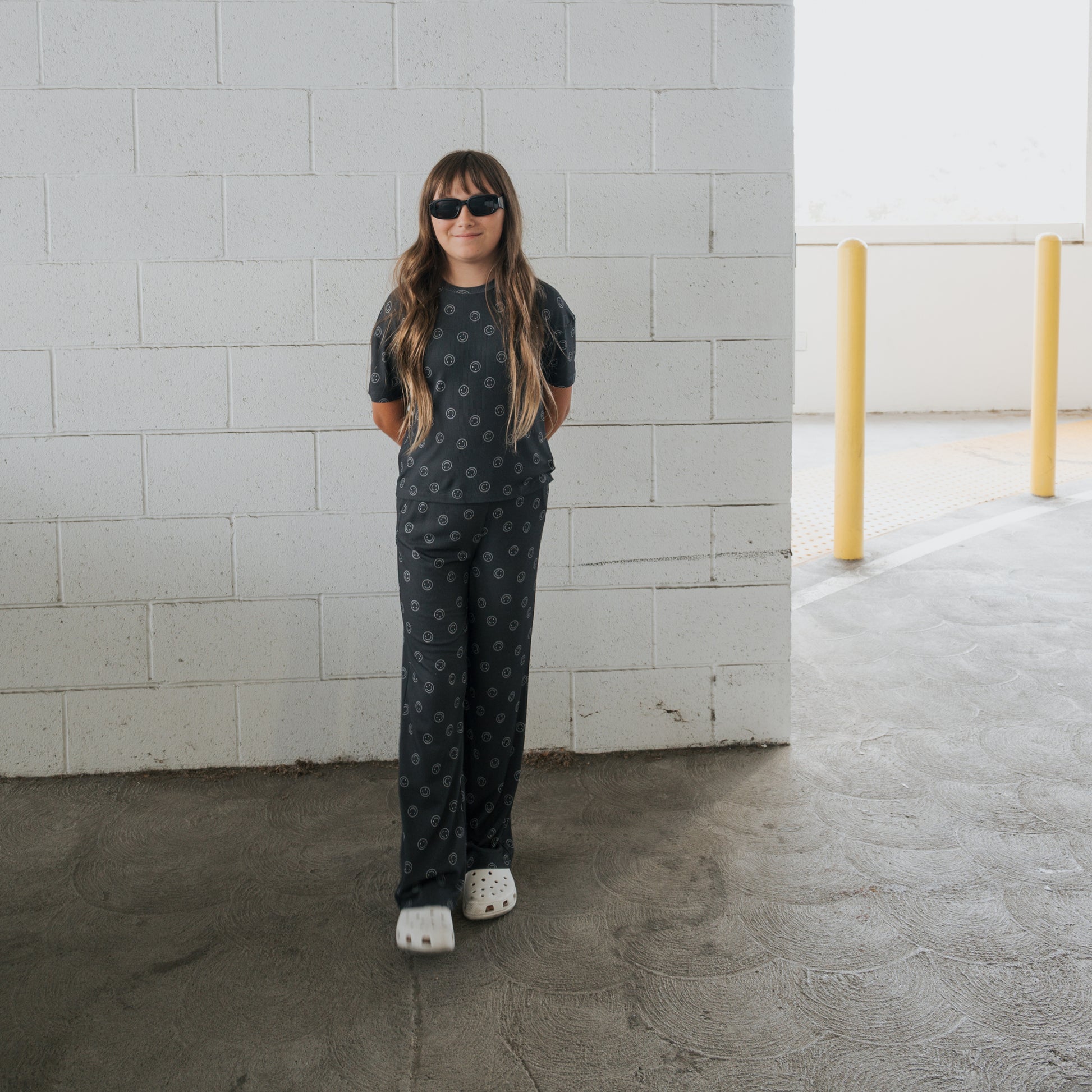 A young person in the forever french baby Child Boxy Tee Lounge Set | Smiley Ribbed stands in front of a white brick wall in a parking garage, sporting long hair, black sunglasses, and white clogs with hands behind their back.