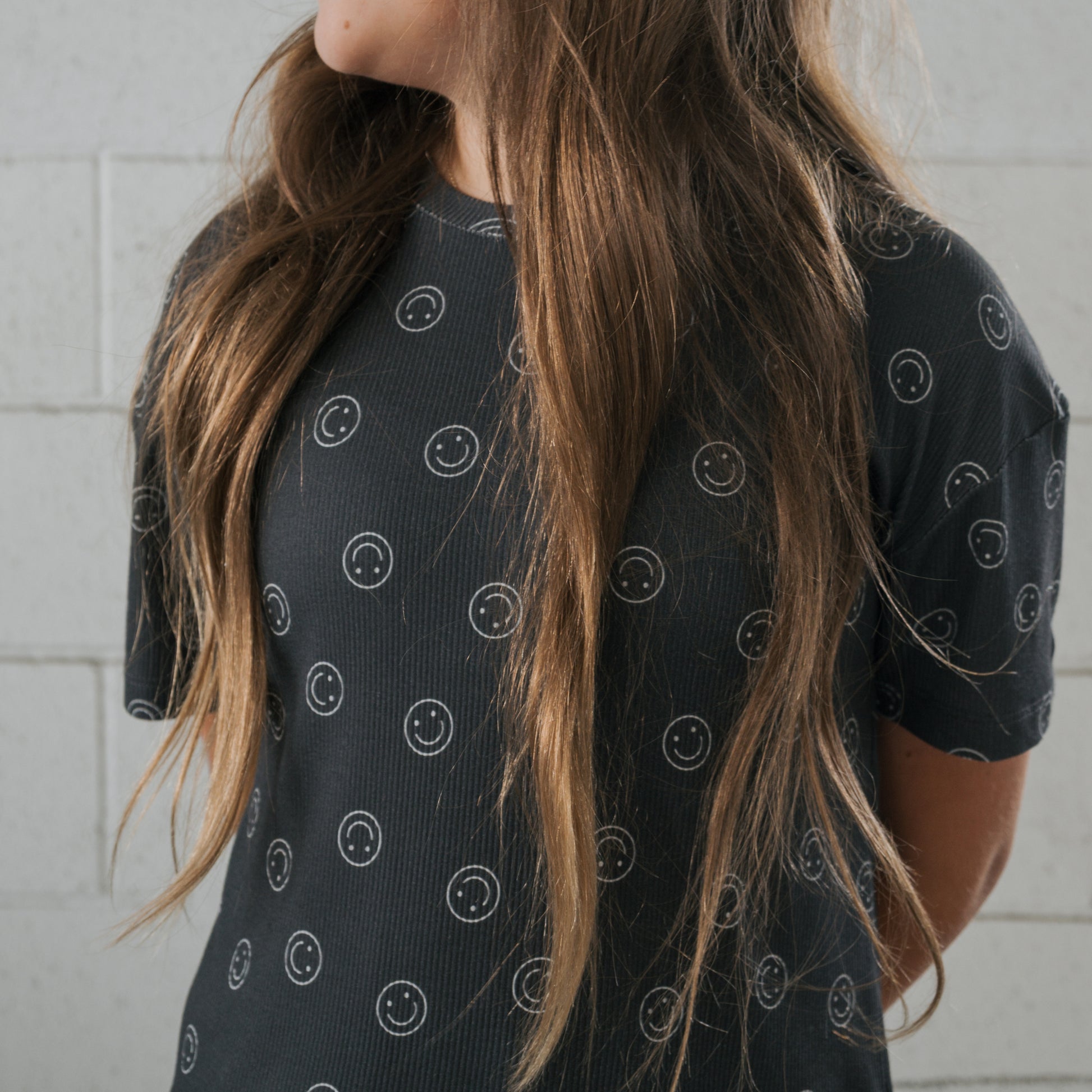 A person with long brown hair wears the forever french baby Child Boxy Tee Lounge Set in Smiley Ribbed, a comfy black bamboo T-shirt with white smiley faces, standing before a light gray brick wall, their face turned away.