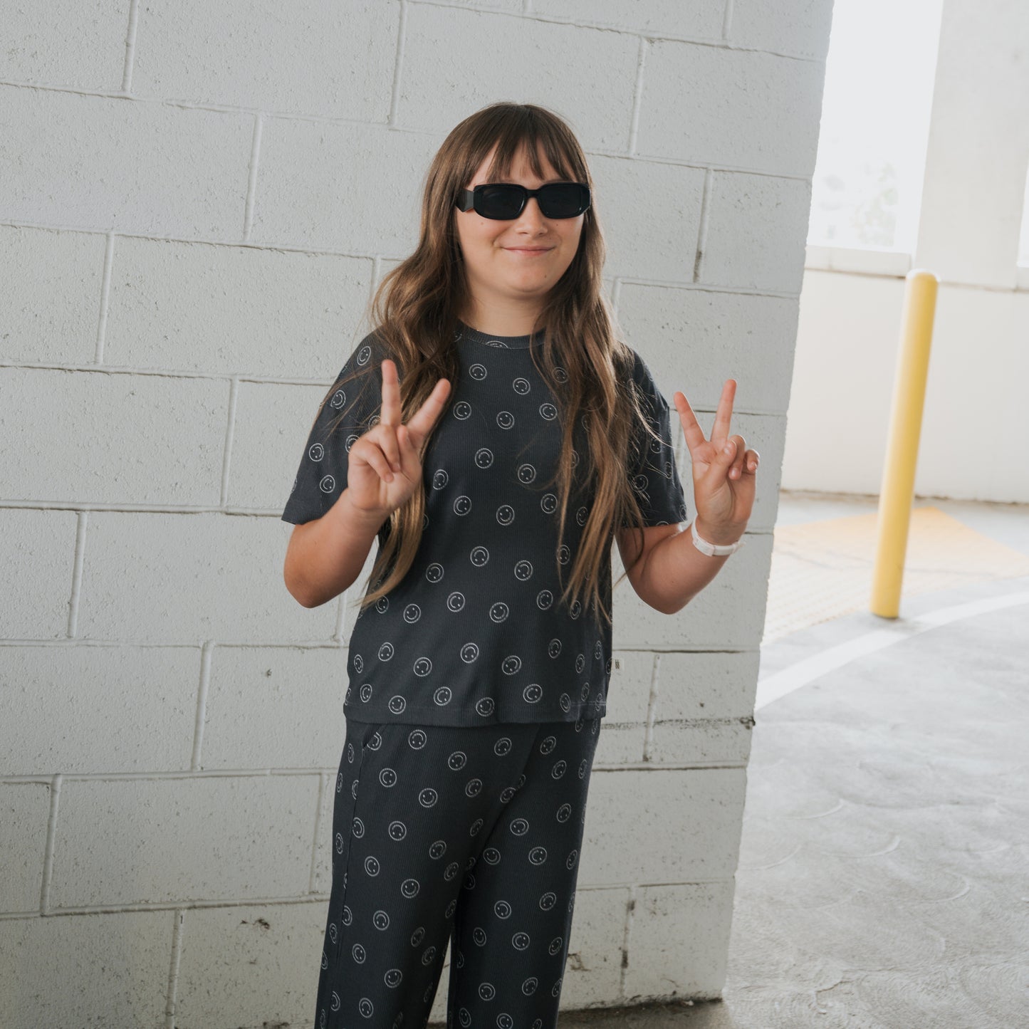 A young person in the forever french baby Child Boxy Tee Lounge Set | Smiley Ribbed, with long hair and sunglasses, stands before a white brick wall, flashing peace signs with both hands.