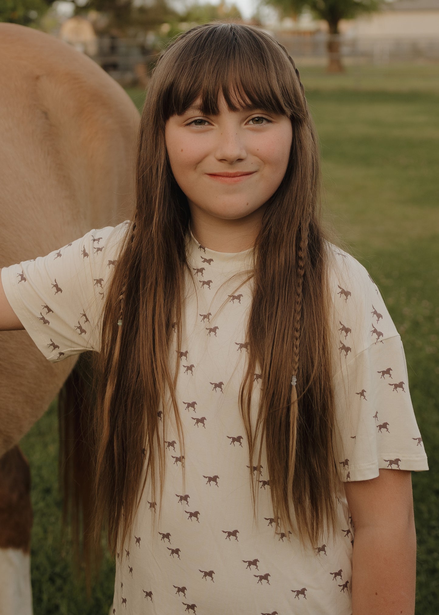 A smiling young girl with a small braid stands outdoors beside a brown horse, wearing the forever french baby Girl's Bamboo Short Set | Wild Horses, surrounded by green grass and trees.