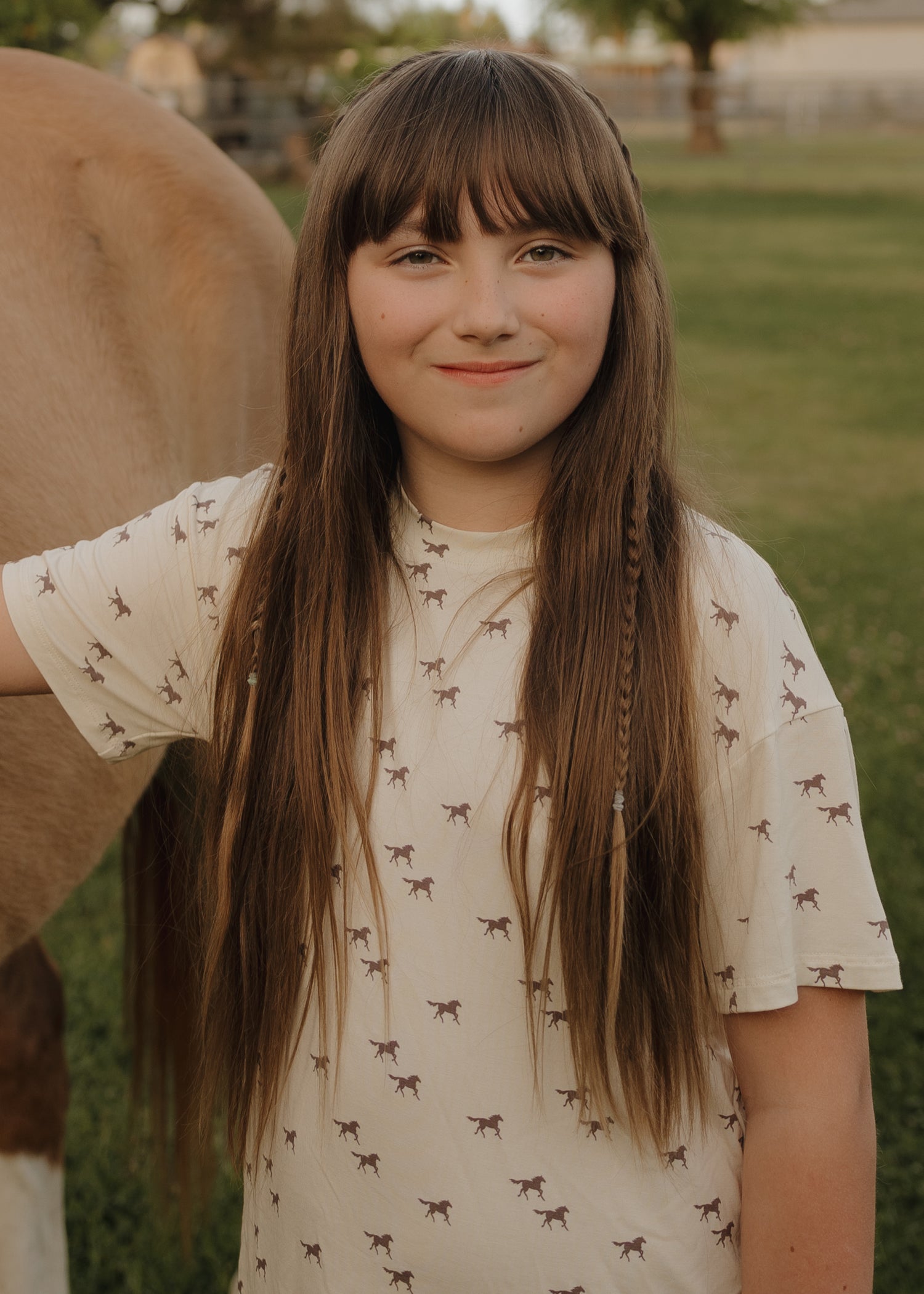 A smiling young girl with a small braid stands outdoors beside a brown horse, wearing the forever french baby Girl's Bamboo Short Set | Wild Horses, surrounded by green grass and trees.
