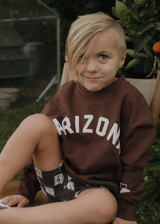 A blonde child sits outdoors on a wooden chair, wearing the forever french baby Child Sweatshirt | ff Arizona and patterned shorts. Surrounded by greenery and an orange tree, this look is perfect for casual, matching family outfits.
