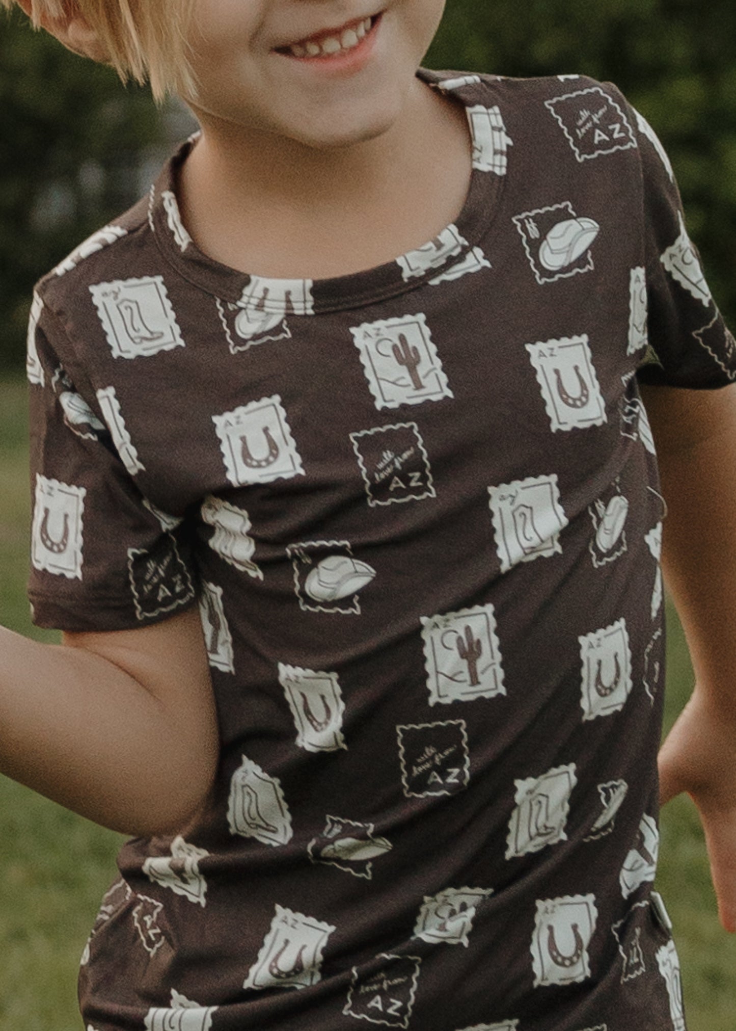 A child smiles outdoors in the forever french baby Bamboo Short Set | With Love from AZ—a brown T-shirt featuring white postage stamp illustrations of cacti, cowboy hats, horseshoes, and "AZ" letters.