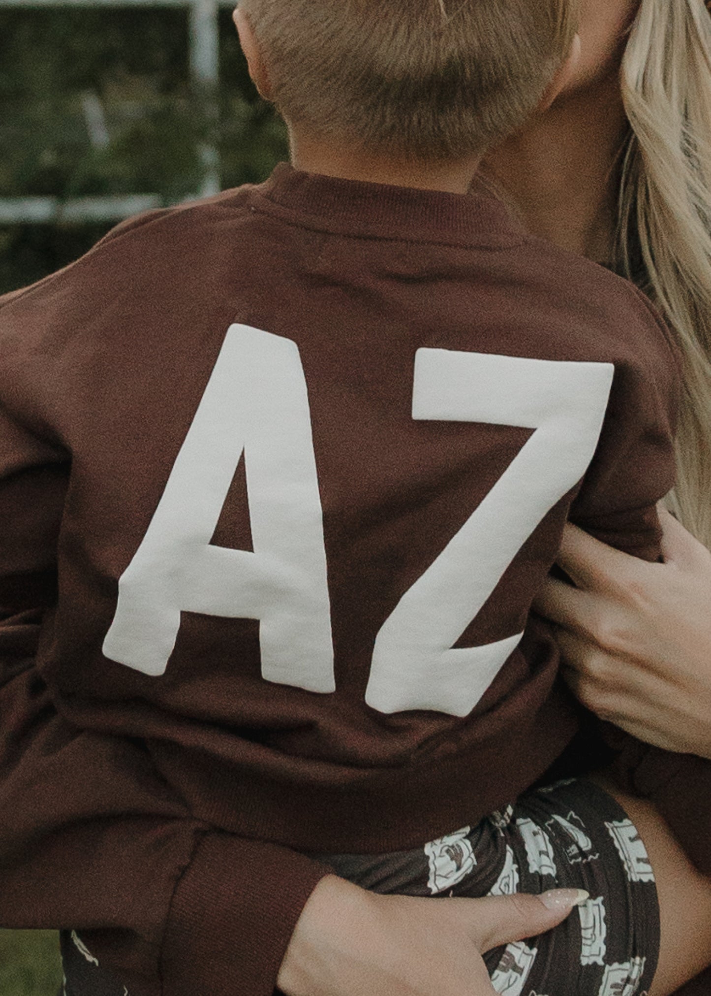 A child wearing the forever french baby Child Sweatshirt | ff Arizona, featuring bold white letters on the back, is being held by an adult with a partially visible face.