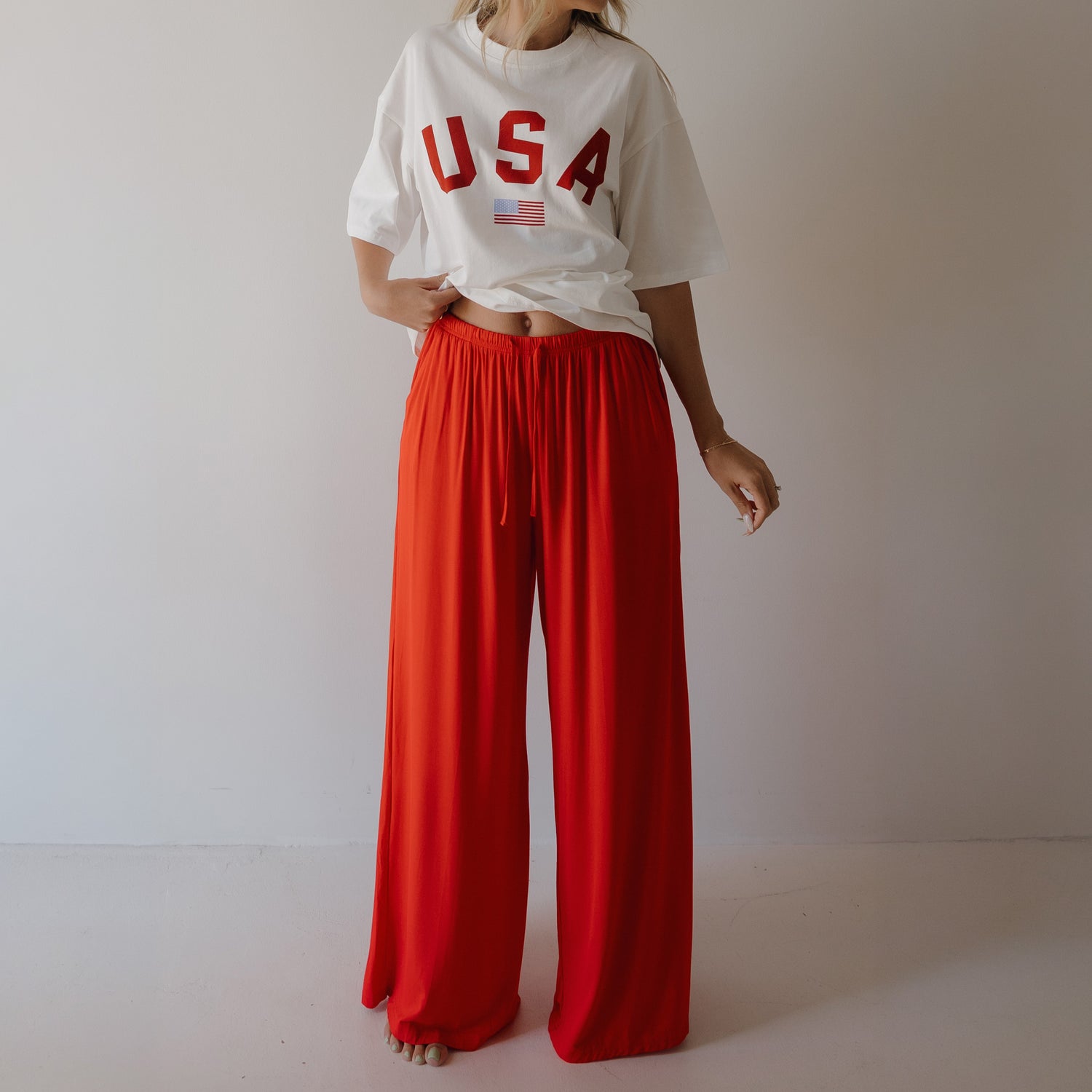 A person stands against a plain, light background wearing a loose white "USA" tee and forever french baby Women's Bamboo Wide Leg Pants in Cherry Red. The pants are high-waisted and made from breathable bamboo fabric. The person's face is not visible.