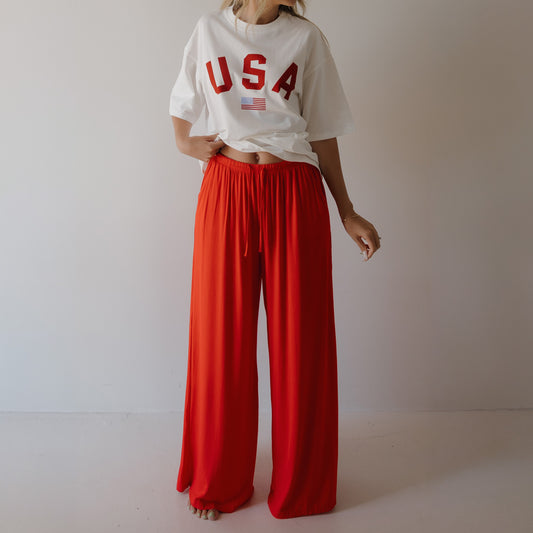 A person stands against a plain, light background wearing a loose white "USA" tee and forever french baby Women's Bamboo Wide Leg Pants in Cherry Red. The pants are high-waisted and made from breathable bamboo fabric. The person's face is not visible.
