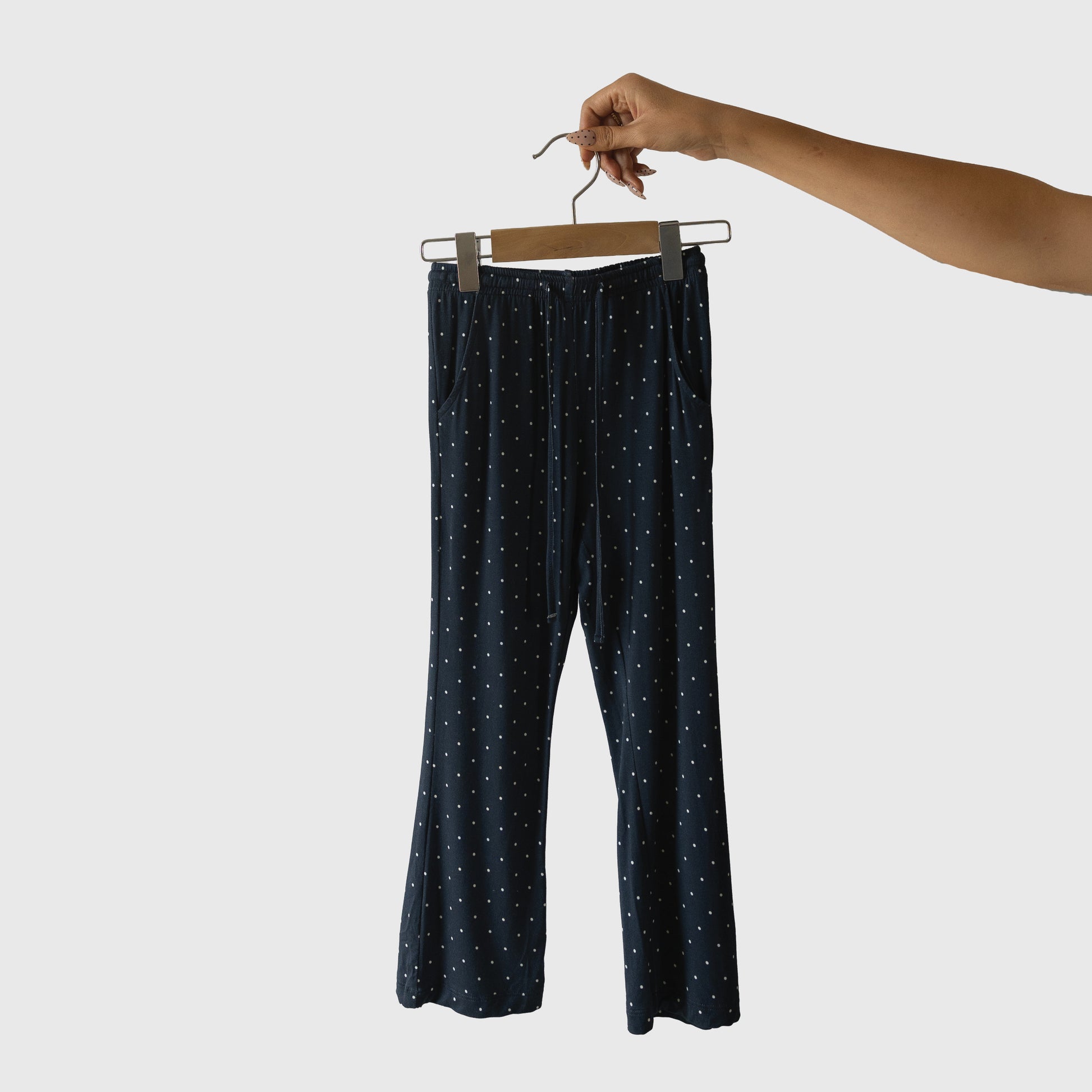 A hand holds a hanger with navy blue pajama pants featuring white polka dots from the forever french baby Child Short Sleeve Bamboo Lounge Set | Emery, part of a hypo-allergenic collection, isolated on a plain white background.
