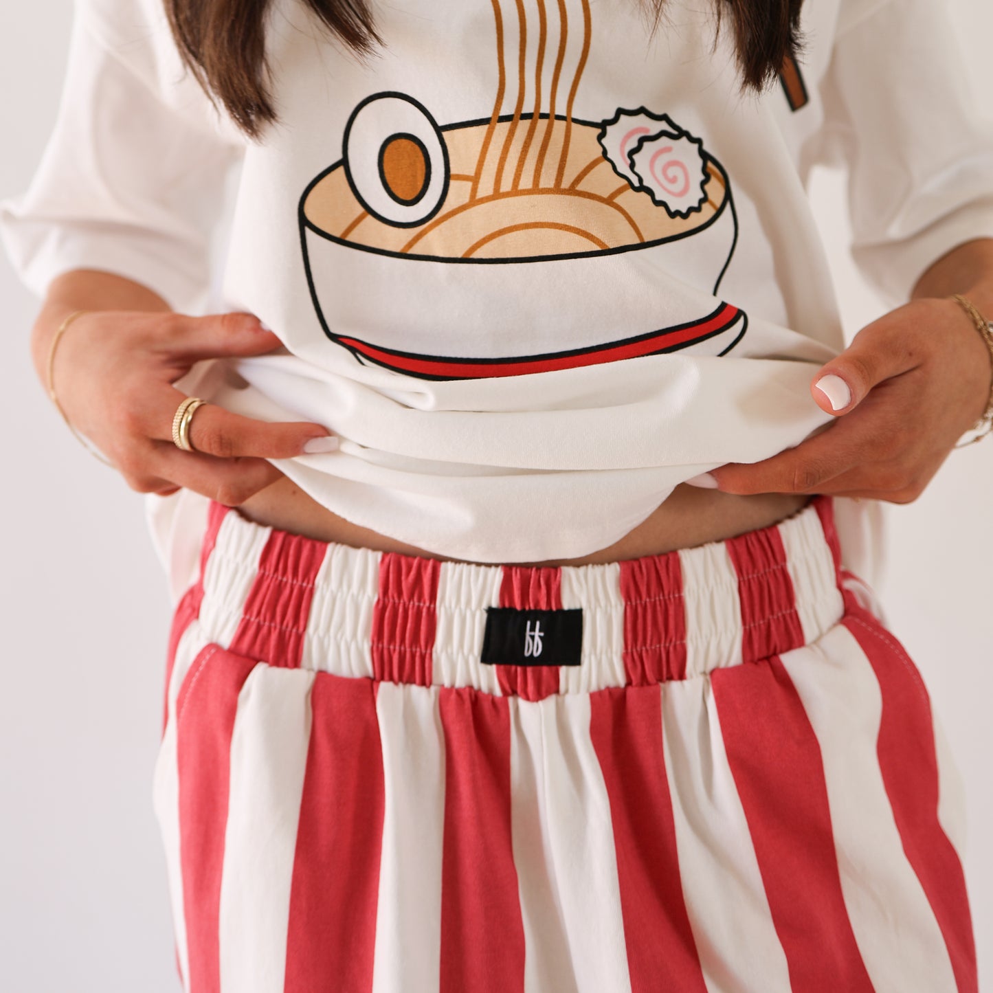 A person lifts their white shirt with a cartoon ramen bowl, revealing forever french baby Women's Wide Leg Pants | Tokyo Stripe. Their nails are light pink, and they accessorize with multiple rings and a bracelet.