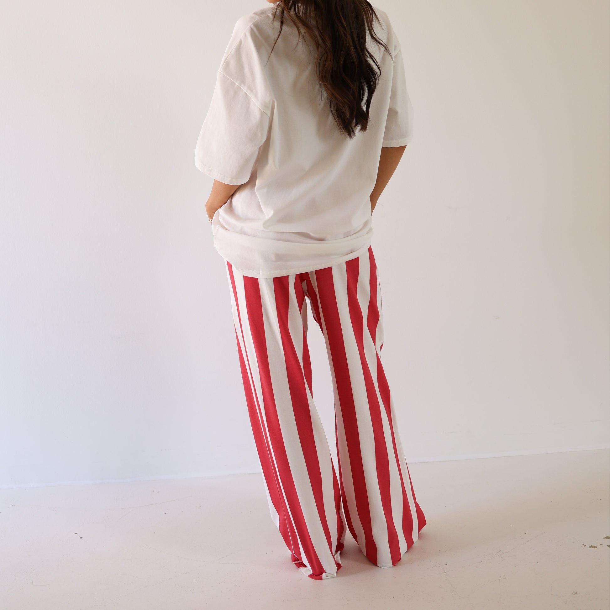 A person faces away, wearing a loose white T-shirt and forever french baby Women's Wide Leg Pants | Tokyo Stripe, featuring red and white vertical stripes with an elastic waistband, against a plain white background.