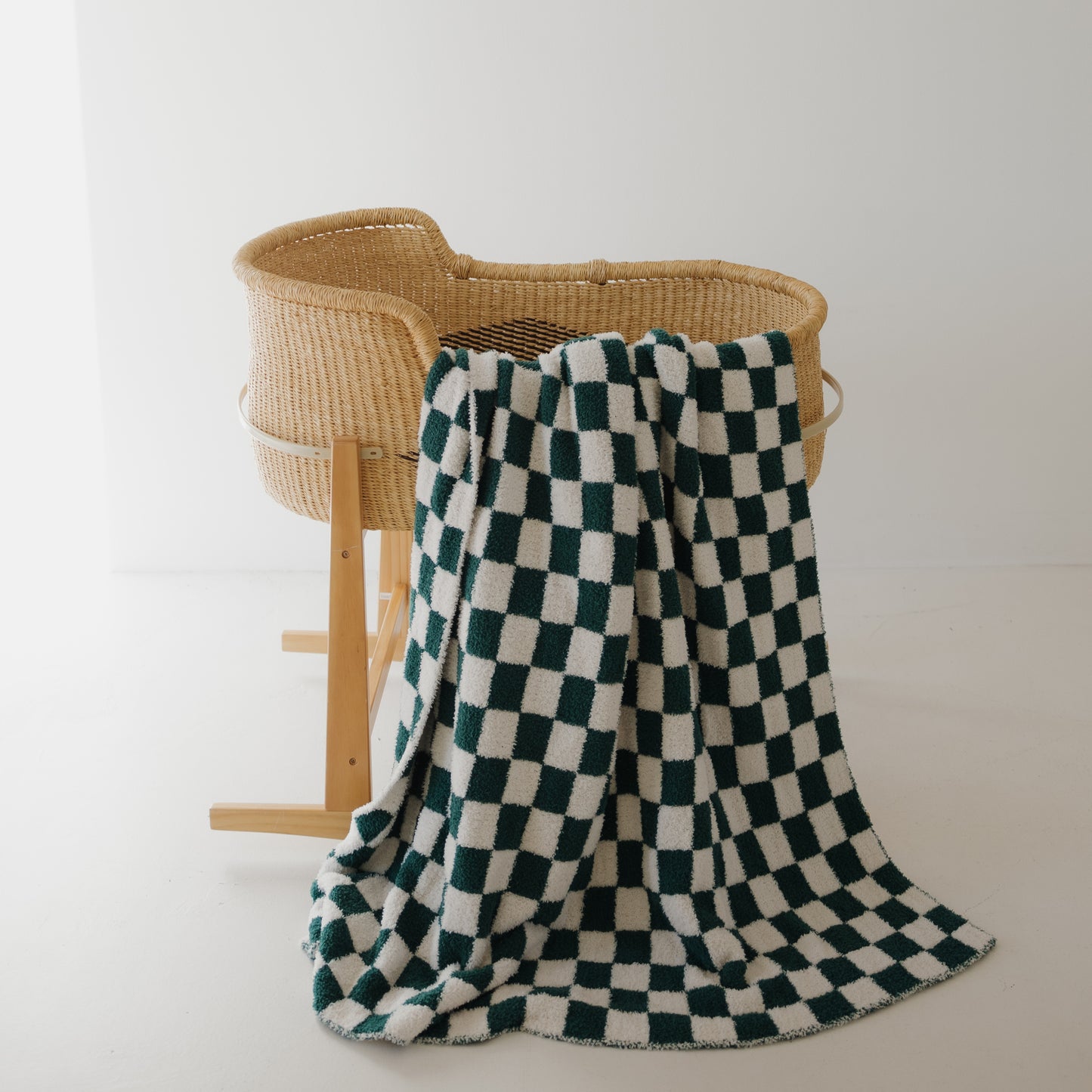 A wicker baby bassinet on wooden legs stands against a white wall, with the forever french baby Plush Blanket | Lucky Micro Check draped over its side and cascading onto the floor.