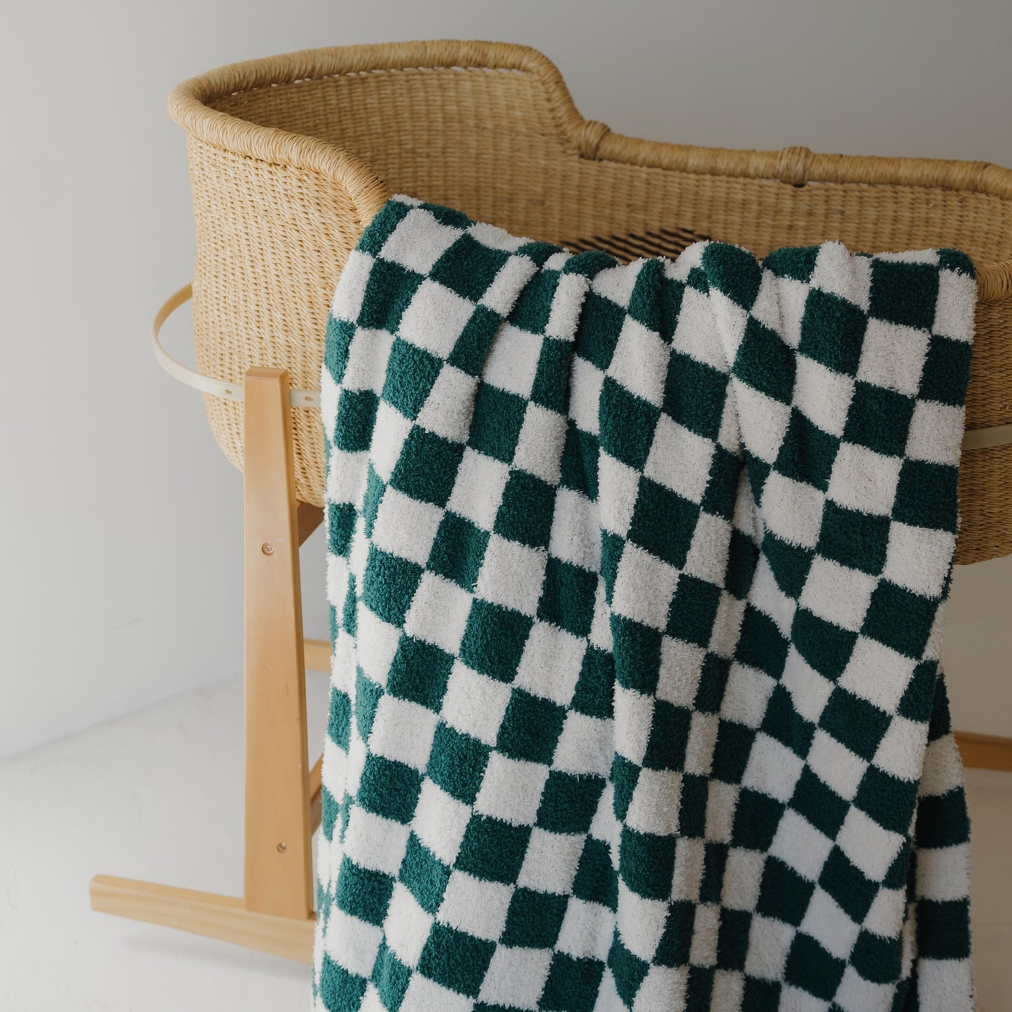 A wicker baby bassinet on wooden legs features the forever french baby Plush Blanket | Lucky Micro Check, Oeko-Tex certified in a green and white checkerboard pattern, draped over its side in a minimal, bright setting.