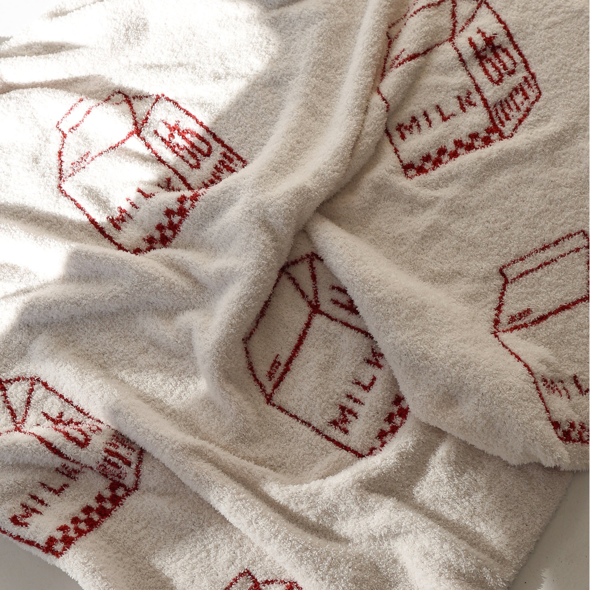 The Plush Blanket | Milk by forever french baby is a soft, cream-colored microfiber feather yarn throw with repeated red "MILK" carton line drawings. Sunlight highlights its cozy, inviting surface.