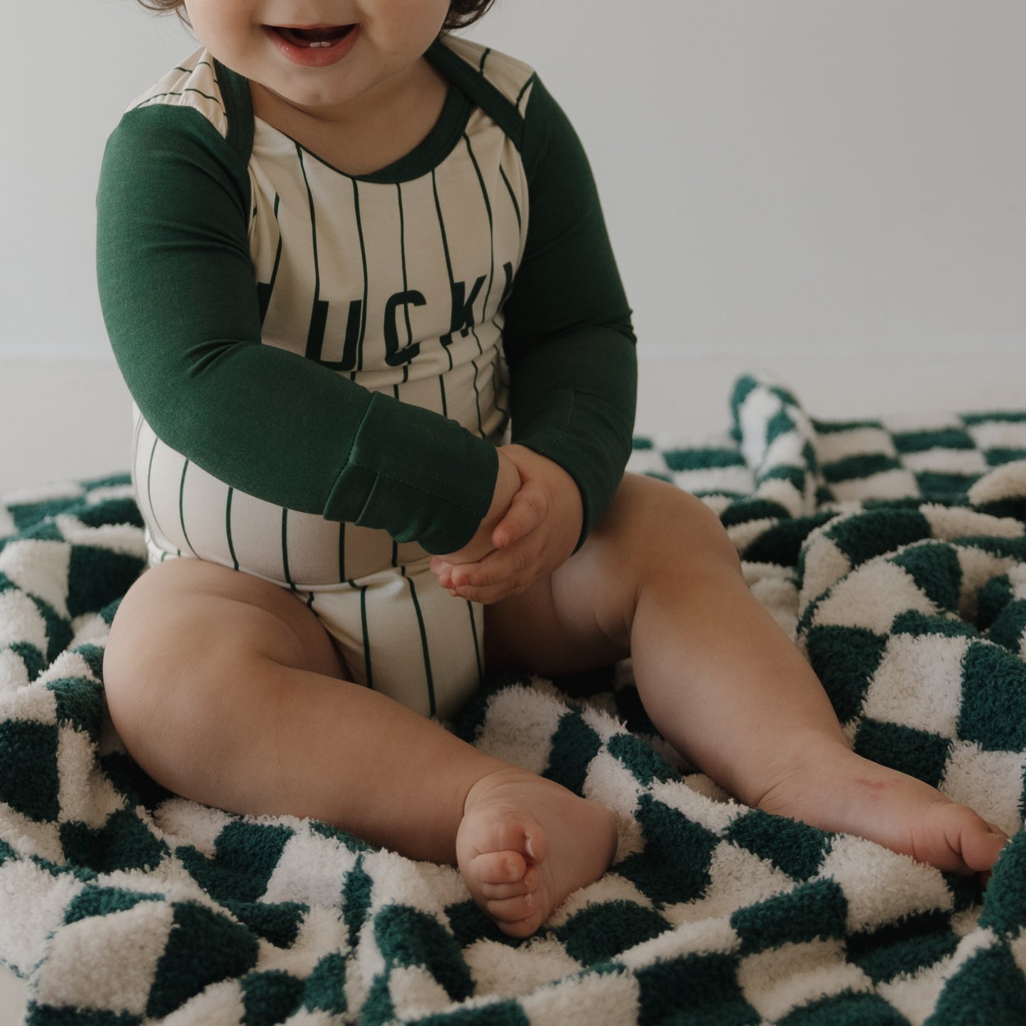 A baby in a green and cream striped onesie sits smiling with crossed feet on the forever french baby Plush Blanket | Lucky Micro Check, an Oeko-Tex certified blanket with a green and white check pattern. The baby's face is partially visible.