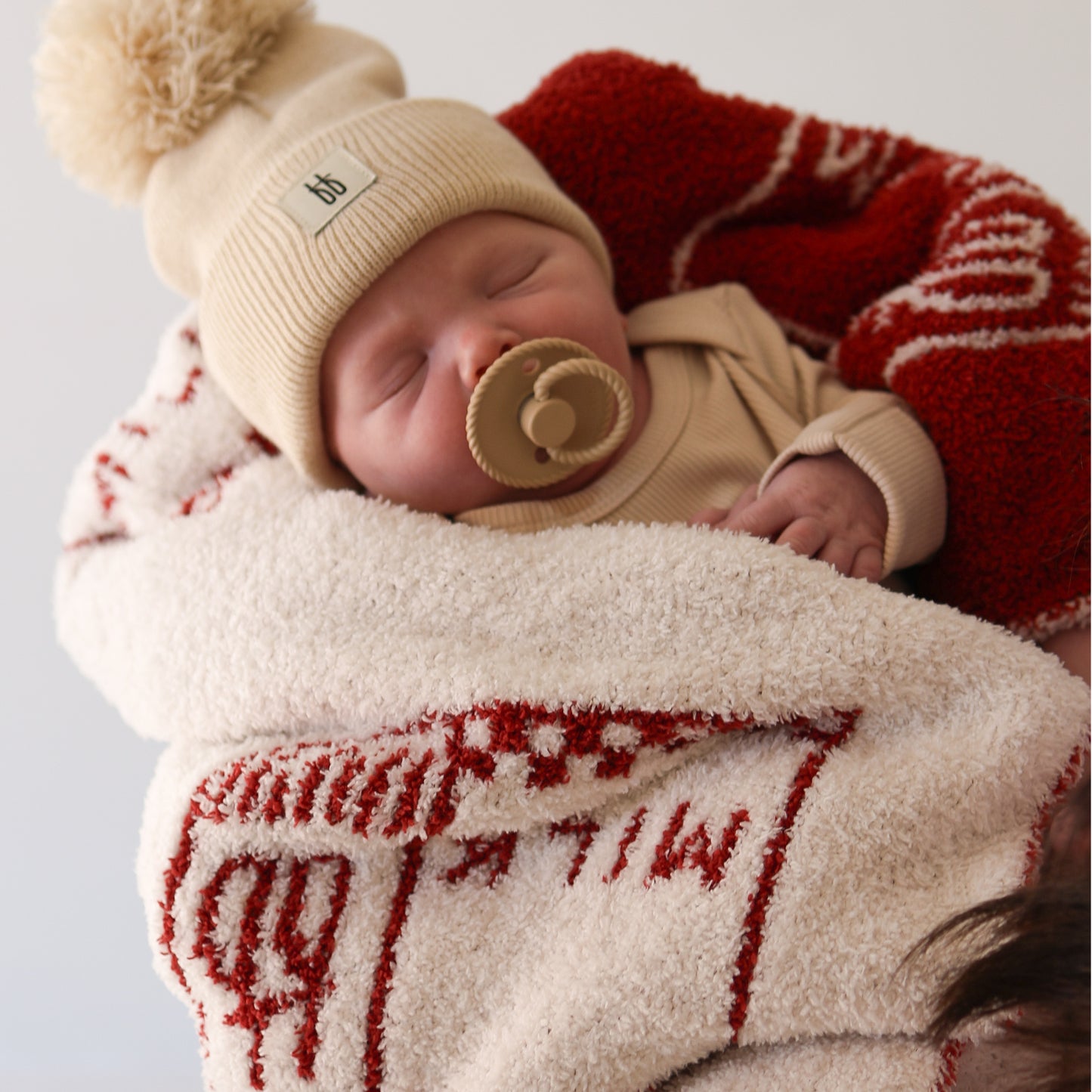 A sleeping baby in a beige outfit with a tan pacifier is wrapped in the forever french baby Plush Blanket | Milk, made of cozy cream and red microfiber feather yarn, keeping them calm, warm, and comfortable.