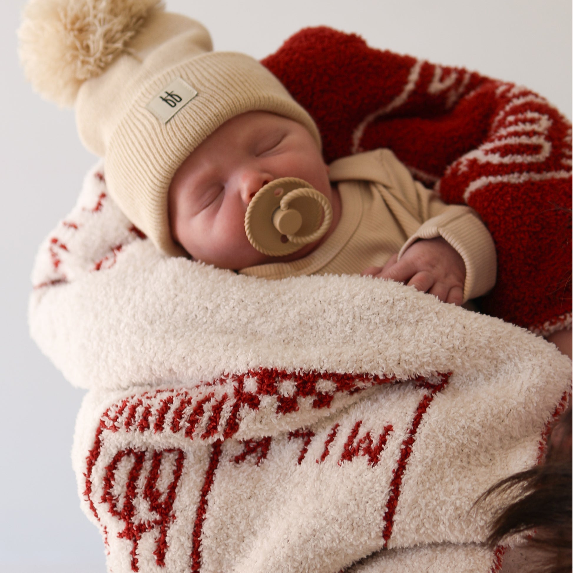 A sleeping baby in a beige outfit with a tan pacifier is wrapped in the forever french baby Plush Blanket | Milk, made of cozy cream and red microfiber feather yarn, keeping them calm, warm, and comfortable.