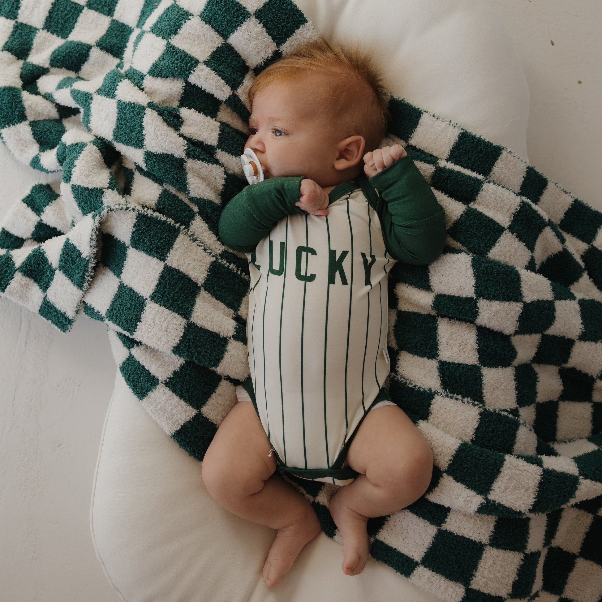 A reddish-haired baby lies on a soft white cushion, wrapped in a green and white checkered blanket, wearing the forever french baby Long Sleeve Bamboo Snapsuit | Lucky and holding a pacifier.