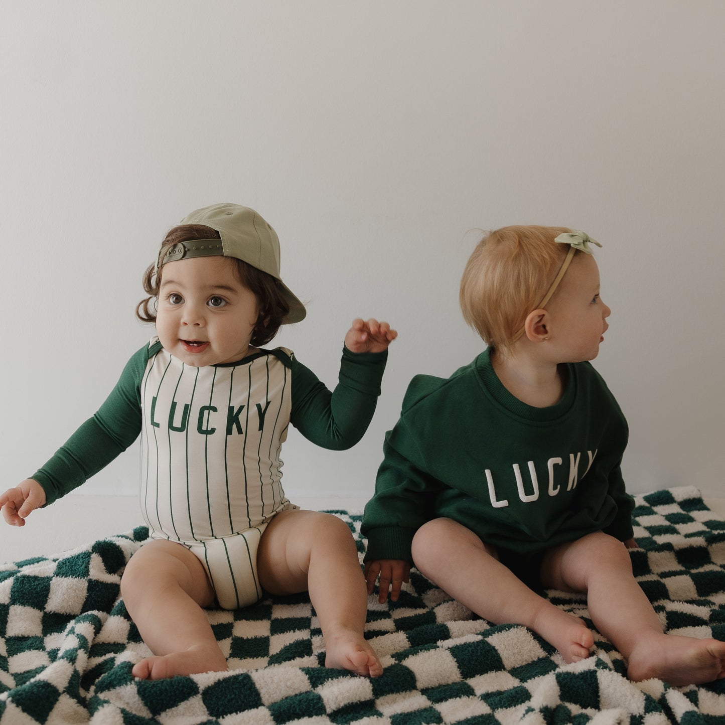Two toddlers sit on a checkered blanket by a white wall. One wears the forever french baby Long Sleeve Bamboo Snapsuit | Lucky and a backwards cap, while the other has on a green "Lucky" sweatshirt with a bow headband, gazing to the side.