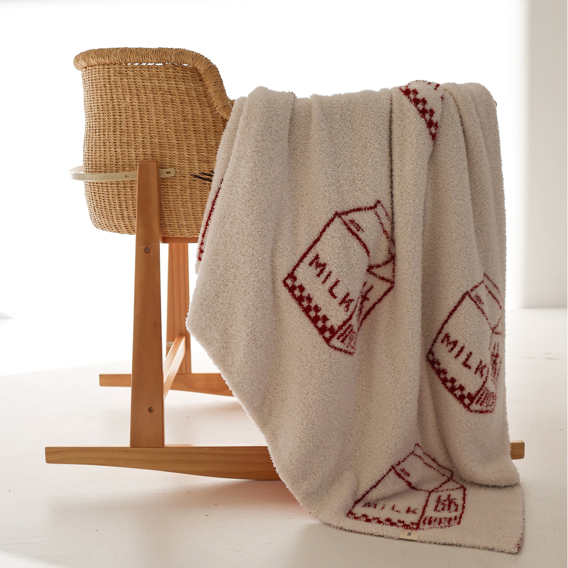 The forever french baby Plush Blanket | Milk, an Oeko-Tex certified cream microfiber feather yarn blanket with red milk carton designs, is draped over a wicker rocking chair in a softly lit, minimal room.