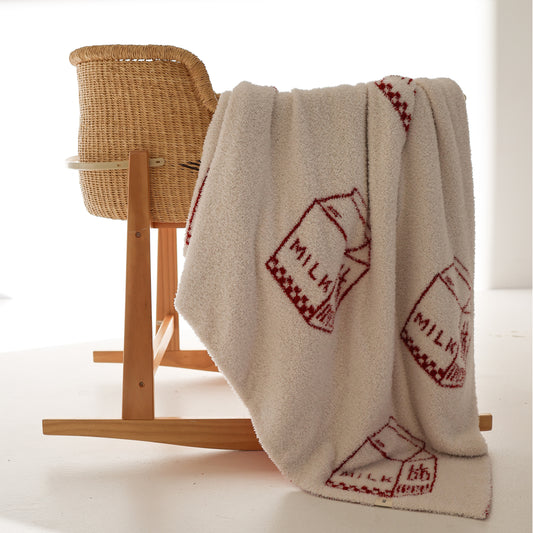 The forever french baby Plush Blanket | Milk, an Oeko-Tex certified cream microfiber feather yarn blanket with red milk carton designs, is draped over a wicker rocking chair in a softly lit, minimal room.