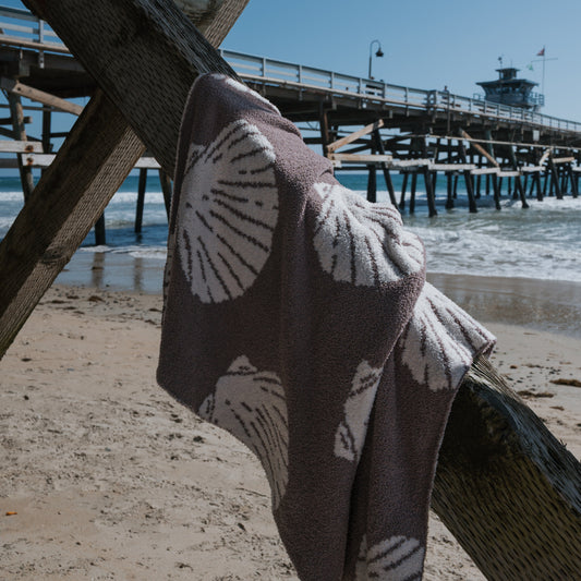 The Plush Blanket | Seashells by forever french baby hangs over a wooden rail on a sandy beach, with ocean waves and a pier in the background beneath a clear blue sky.