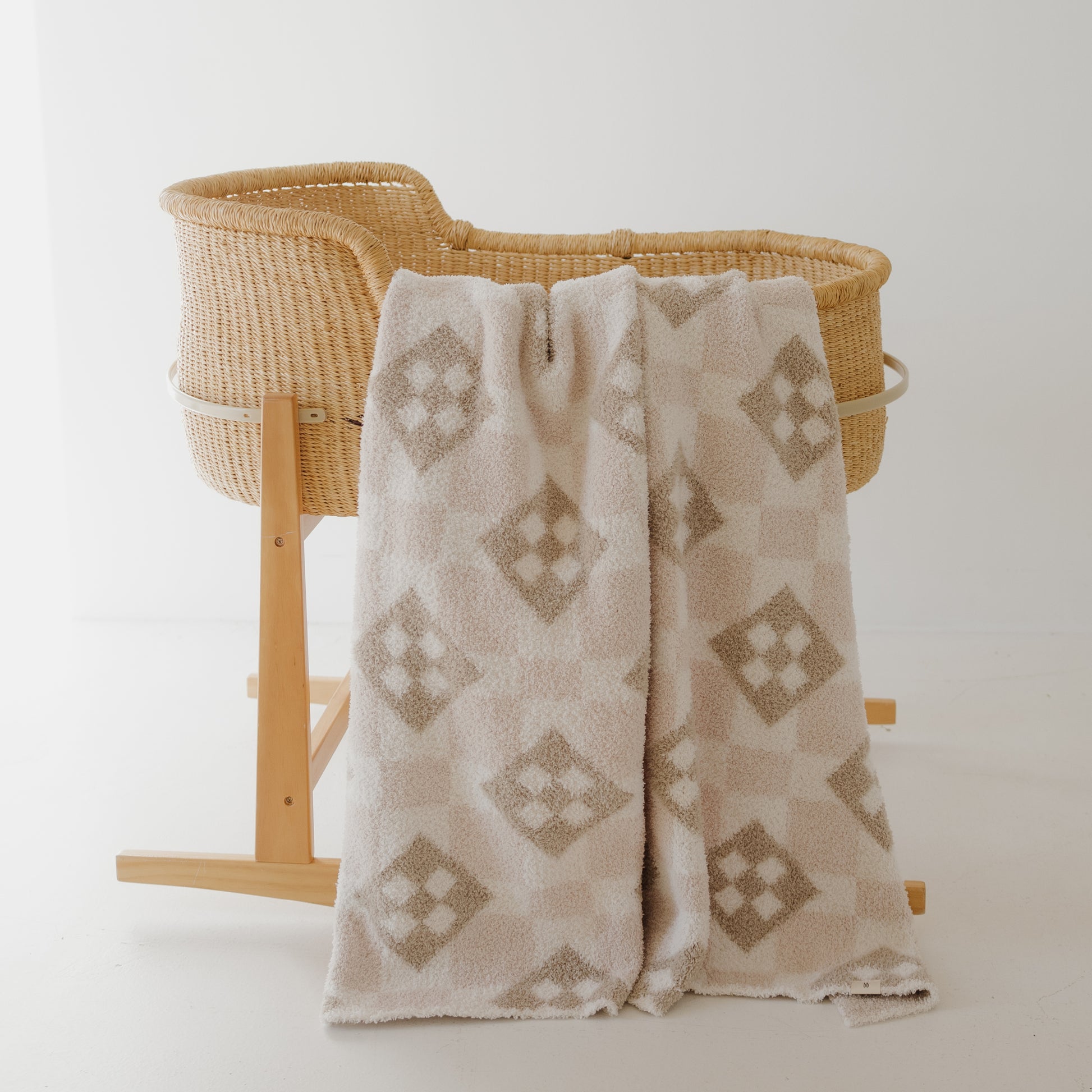 A light brown woven bassinet on wooden legs features a cozy forever french baby Plush Blanket | Cottage Quilt—Oeko-Tex certified in cream and beige pattern—draped over the side, set against a plain white background.