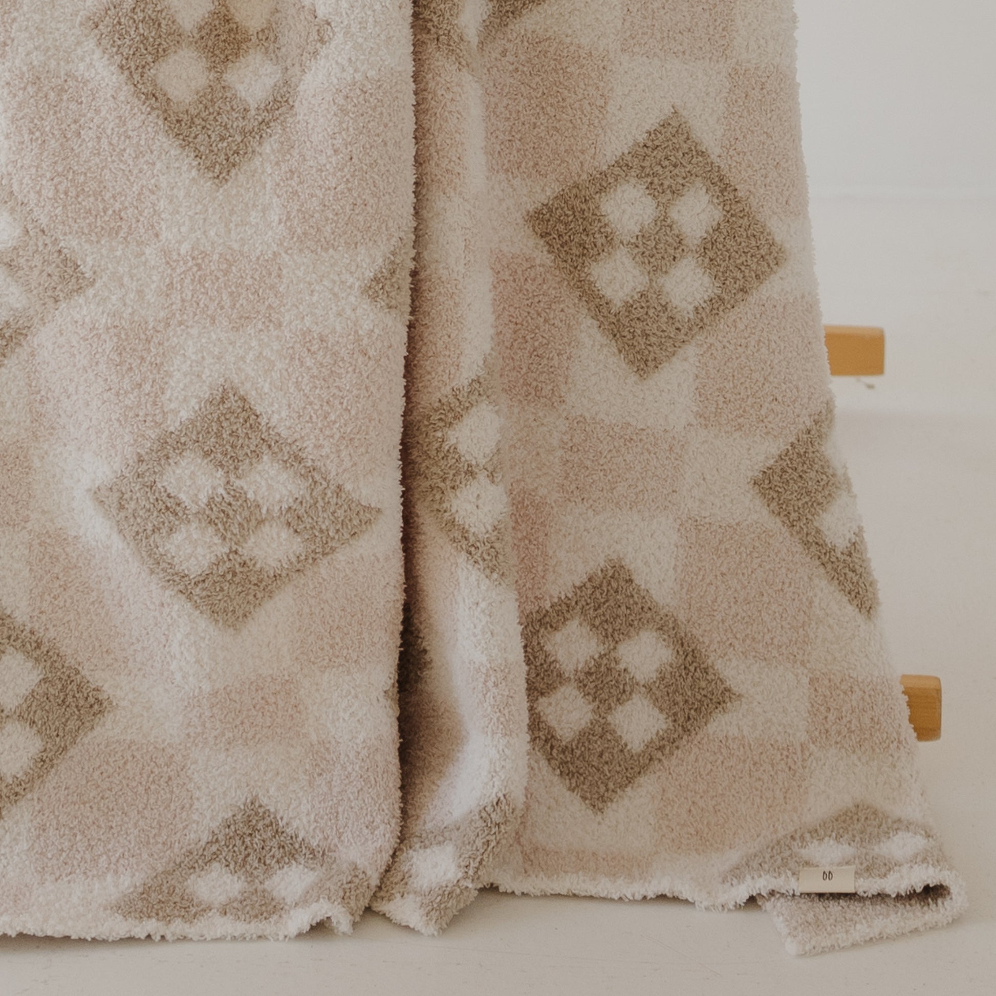 The forever french baby Plush Blanket | Cottage Quilt in beige and cream, with a geometric diamond pattern, drapes over a light wooden stand and partially touches the white floor.