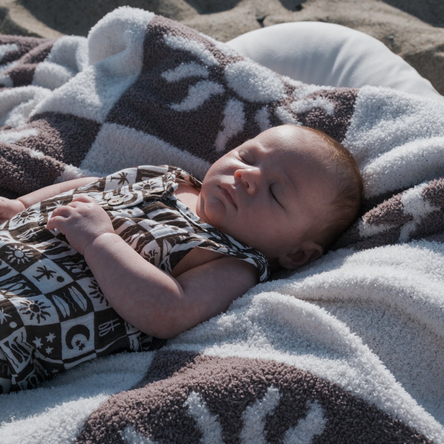 A baby wearing a patterned outfit sleeps peacefully outdoors on a sandy surface, wrapped in the forever french baby Plush Blanket | Summer Sun, which features sun and moon designs and is Oeko-Tex certified.